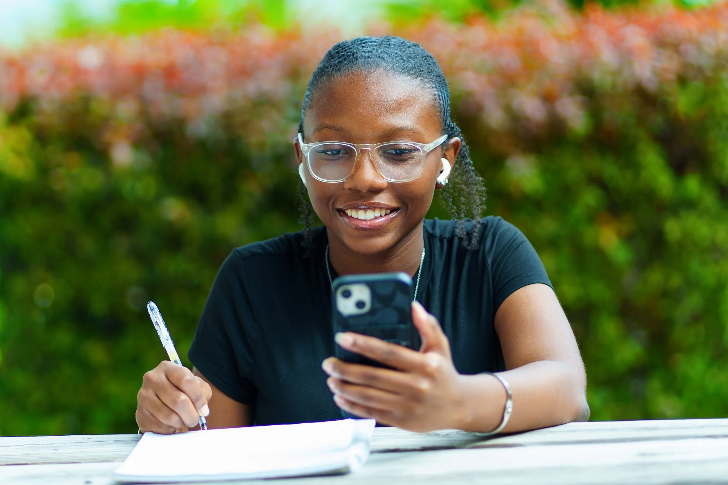 Young woman with glasses and a black shirt sitting at a table outdoors, smiling and looking at her smartphone, with a notebook and pen in her hand, surrounded by blurred greenery.