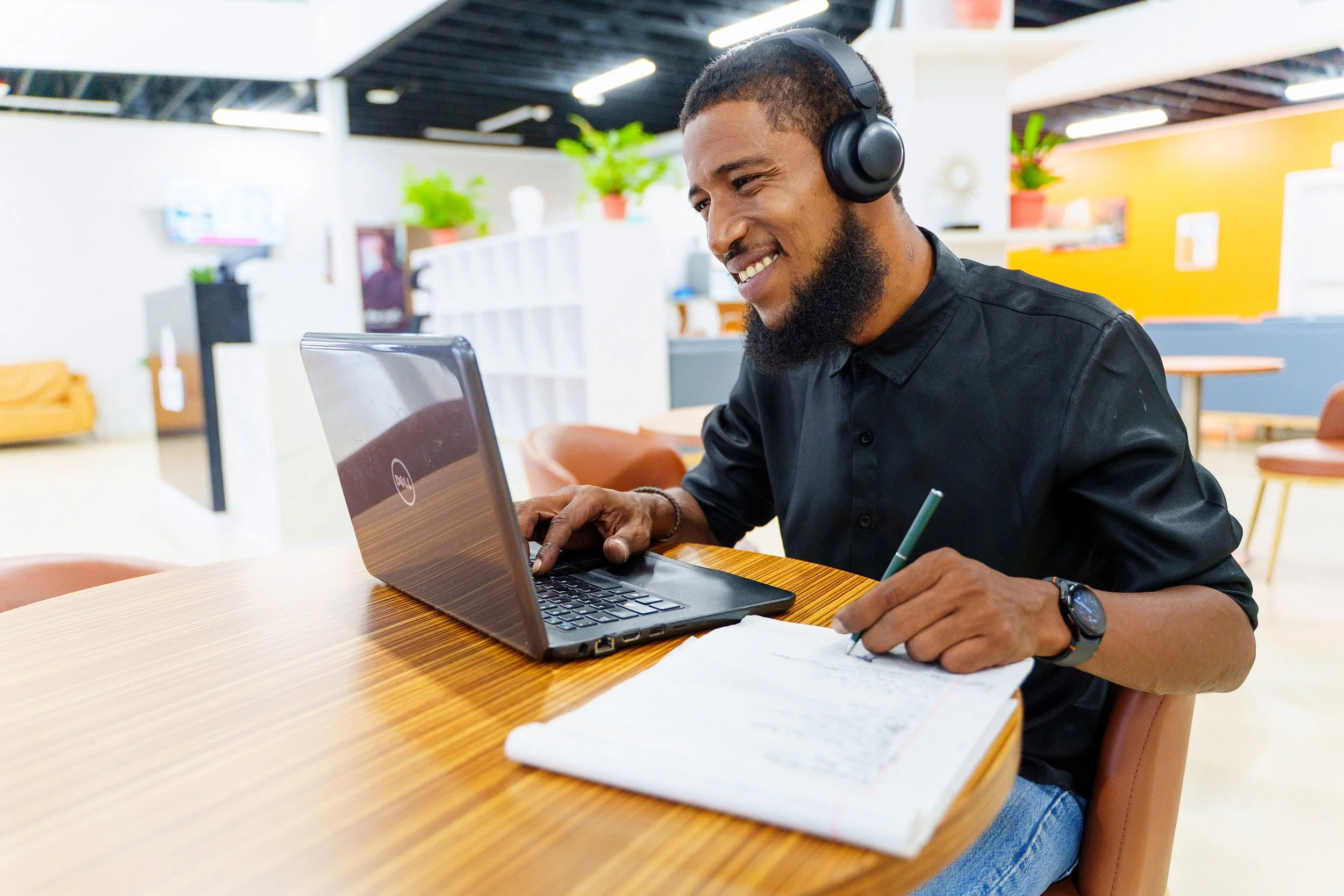 A man with a beard wearing headphones and a black shirt working on a laptop, writing in a notebook at a wooden table in a brightly lit modern office space.