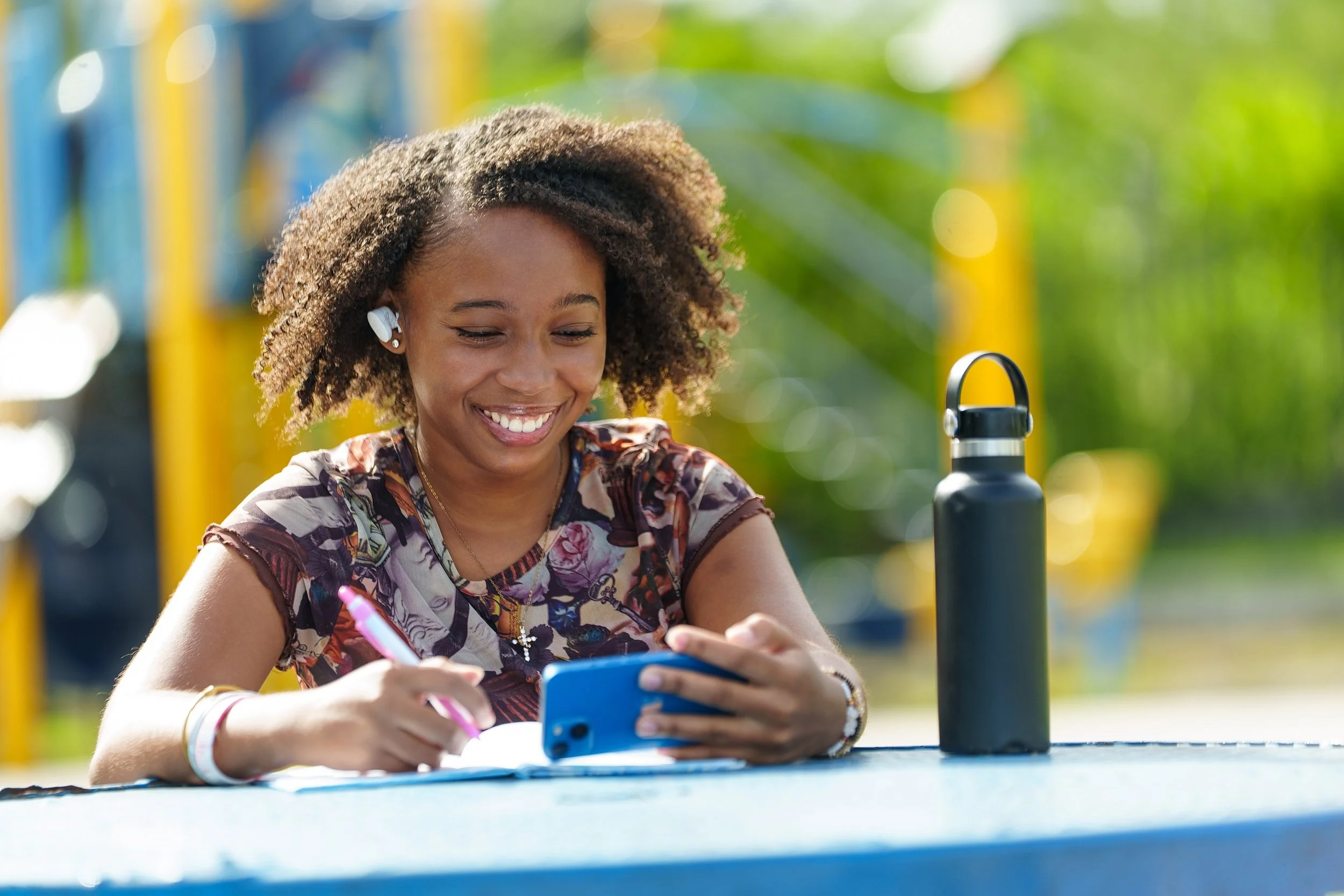 A young woman with curly hair smiling at her phone while sitting at an outdoor table, with a water bottle nearby, during daytime.