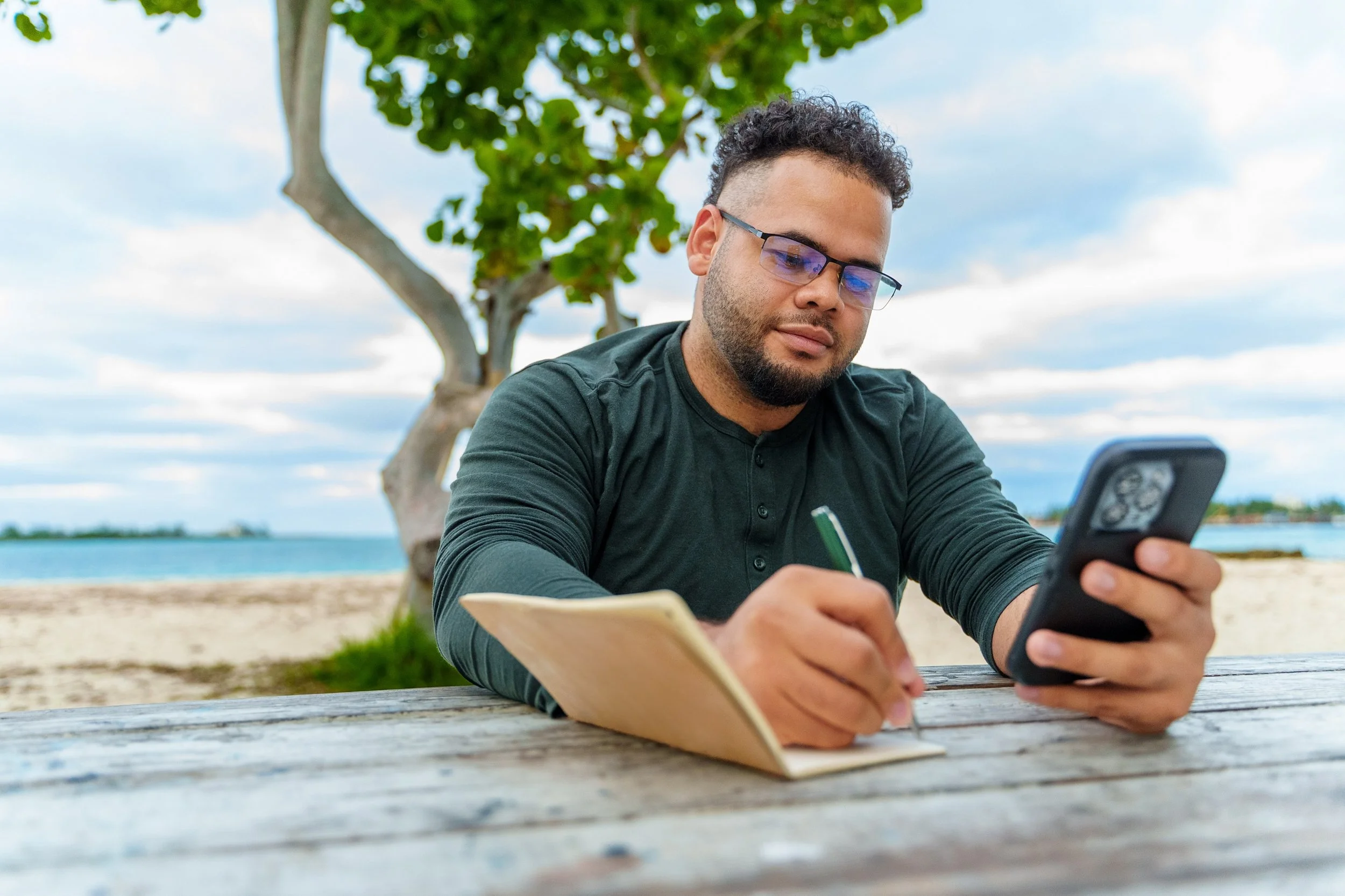 Man sitting at a wooden table outdoors, writing in a notebook with a pen while looking at his smartphone, near a tree and the beach.