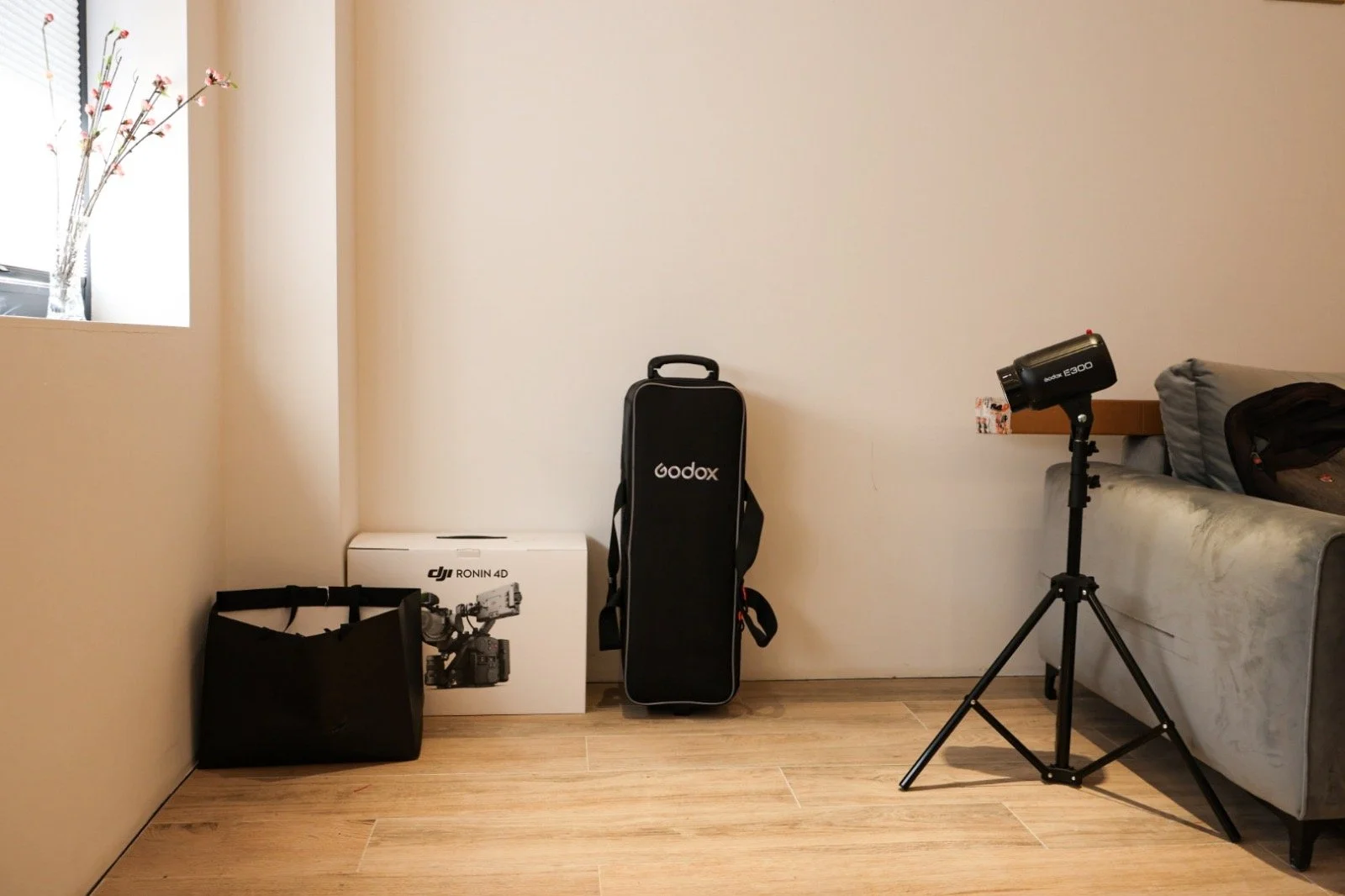 Living room corner with a window on the left, a black 'Godox' tripod case and a DJI Ronin 4D box on the floor, a black bag, and a light on a stand directed at the sofa on the right.