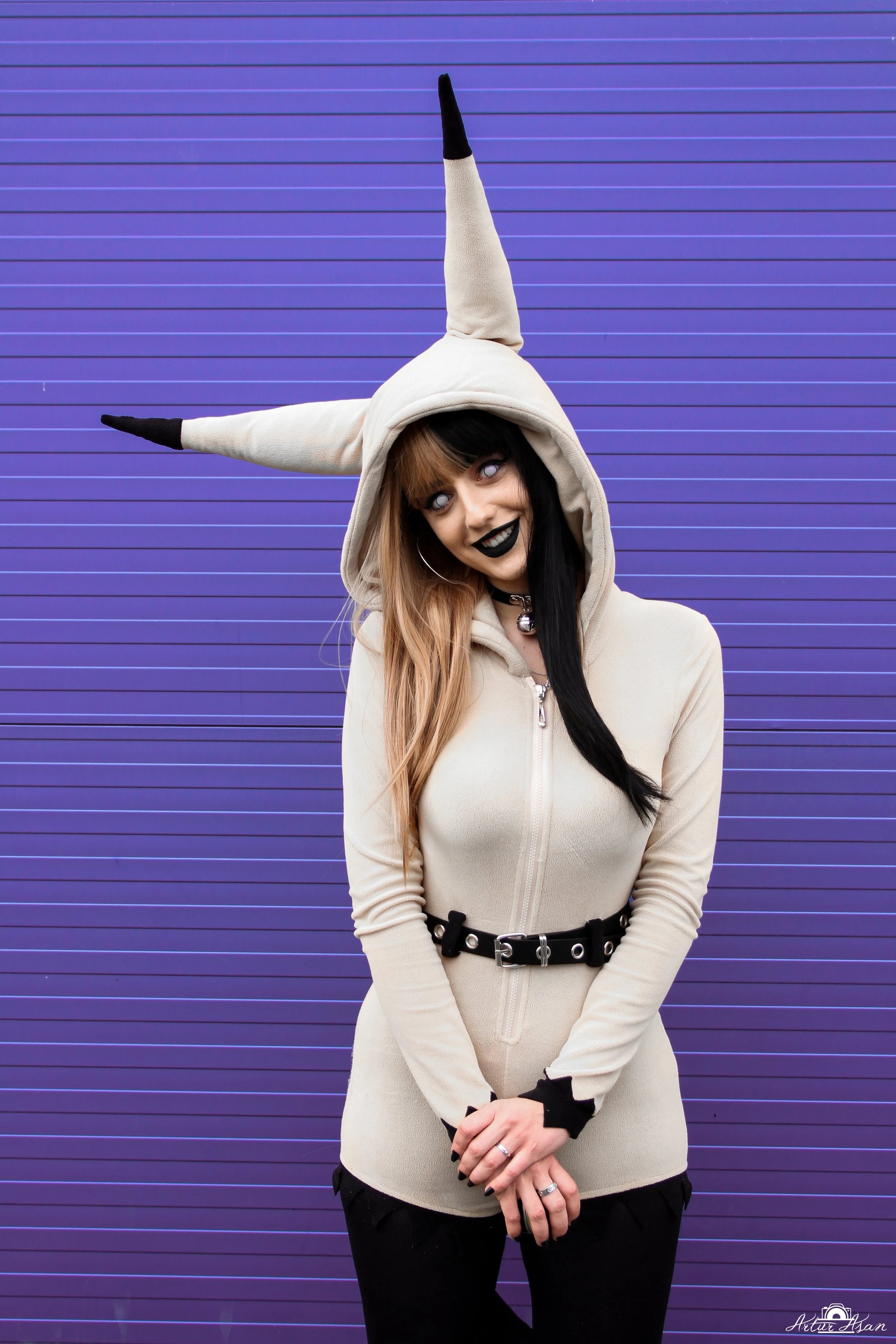 A young woman with multicolored hair wearing a cream-colored hoodie with black accents and a horned hat, standing in front of a purple, horizontally paneled wall.
