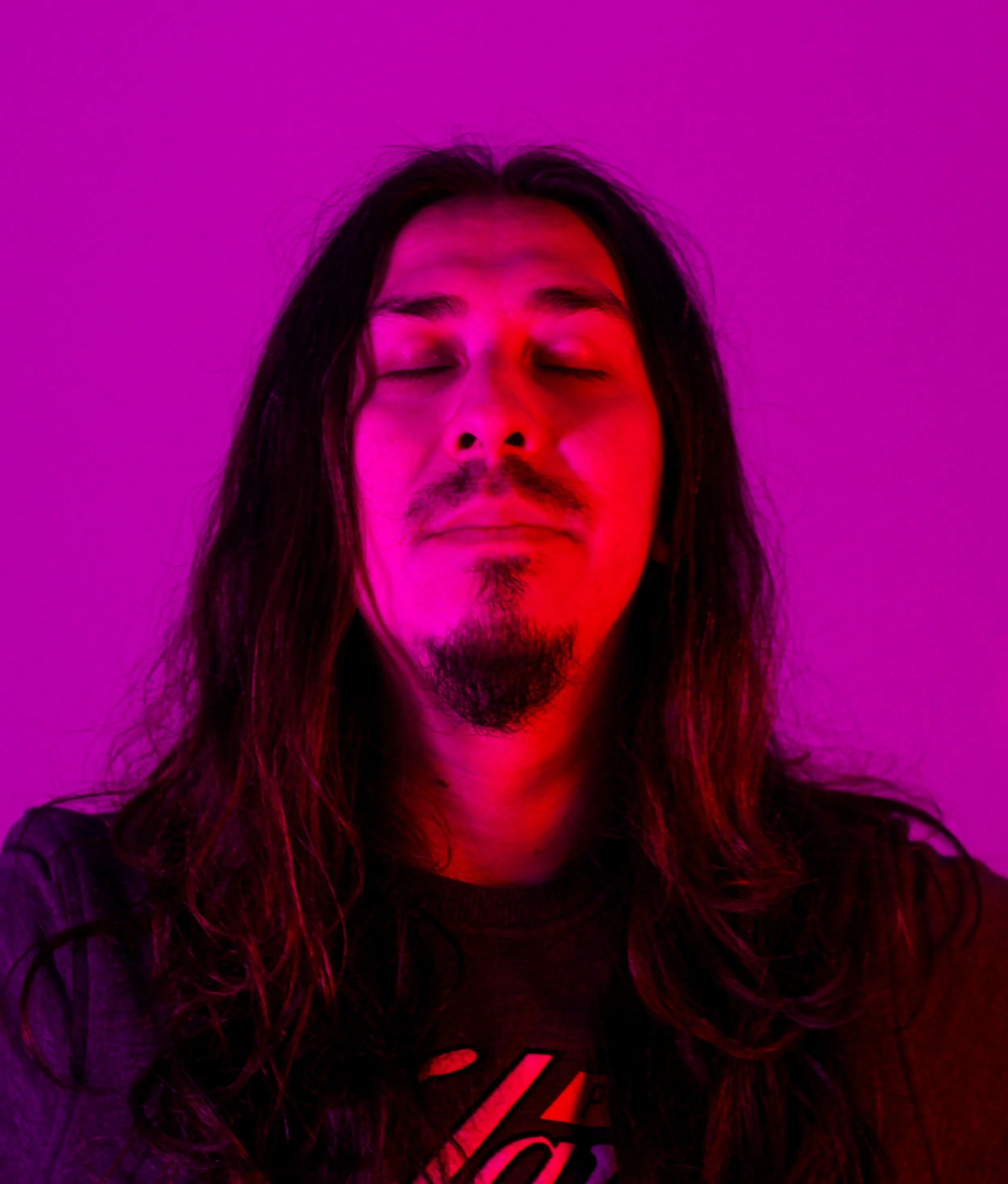 A man with long hair, a mustache, and a goatee, with closed eyes, under pink and red lighting.