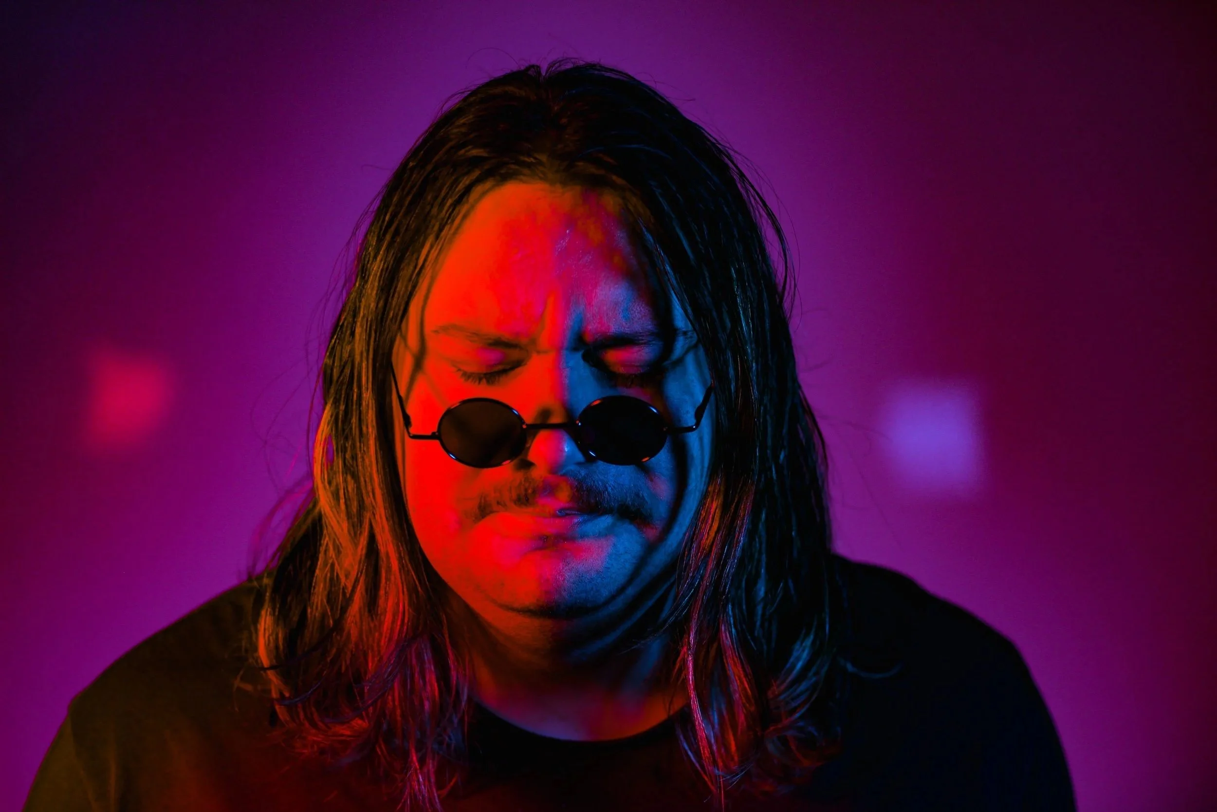 Person with long hair wearing round sunglasses, eyes closed, lit with colorful purple, red, and blue lighting, with a blurred background.