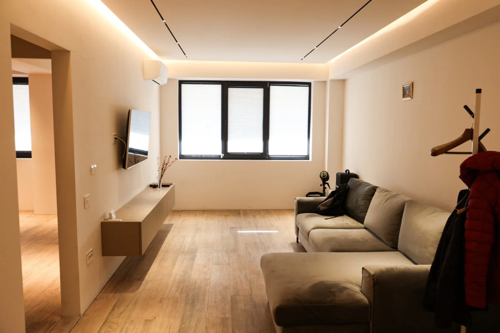 A living room with a beige sectional sofa, a wall-mounted flat-screen TV, a window with blinds, and a light-colored wooden floor.