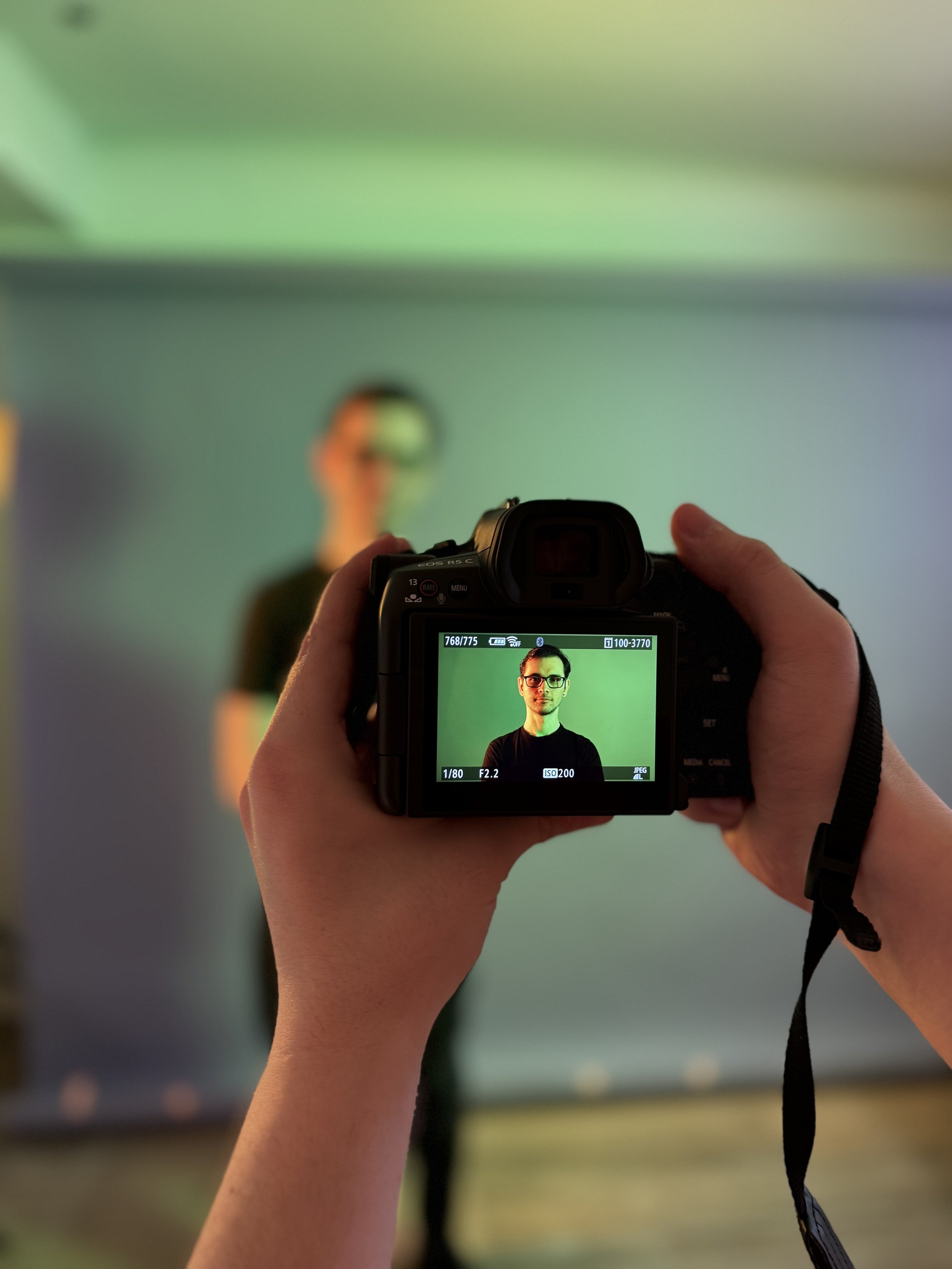 Person takes photographs of a man wearing glasses standing against a green screen background.