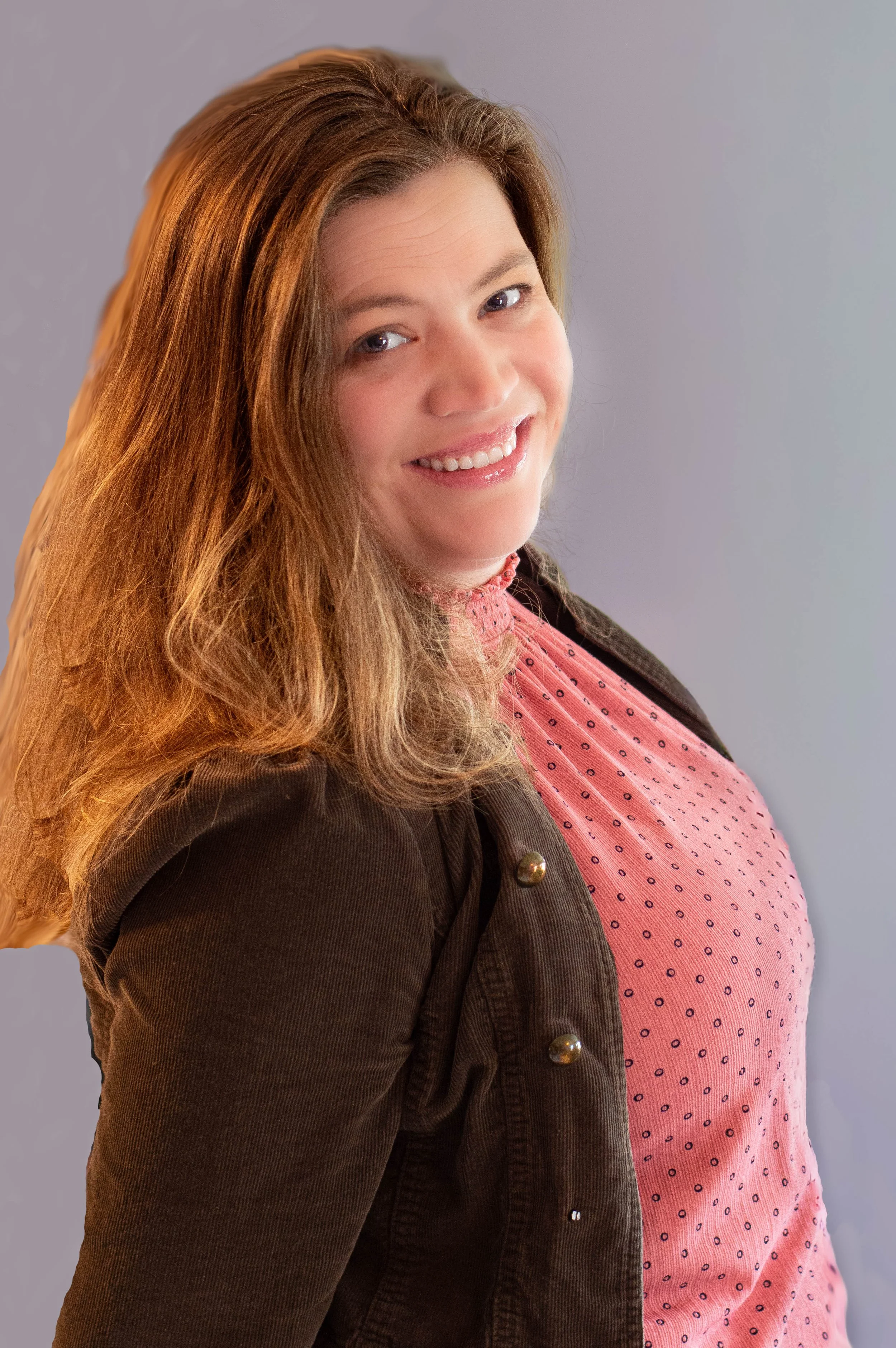 A woman with long, wavy reddish-brown hair smiling, wearing a pink top with small dark patterns and a dark brown blazer with gold buttons, standing against a plain light gray background.