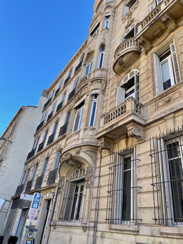 Immeuble ancien en pierre avec fenêtres à volets, balcons en fer forgé, et une signalisation de stationnement à Paris, sous un ciel bleu.