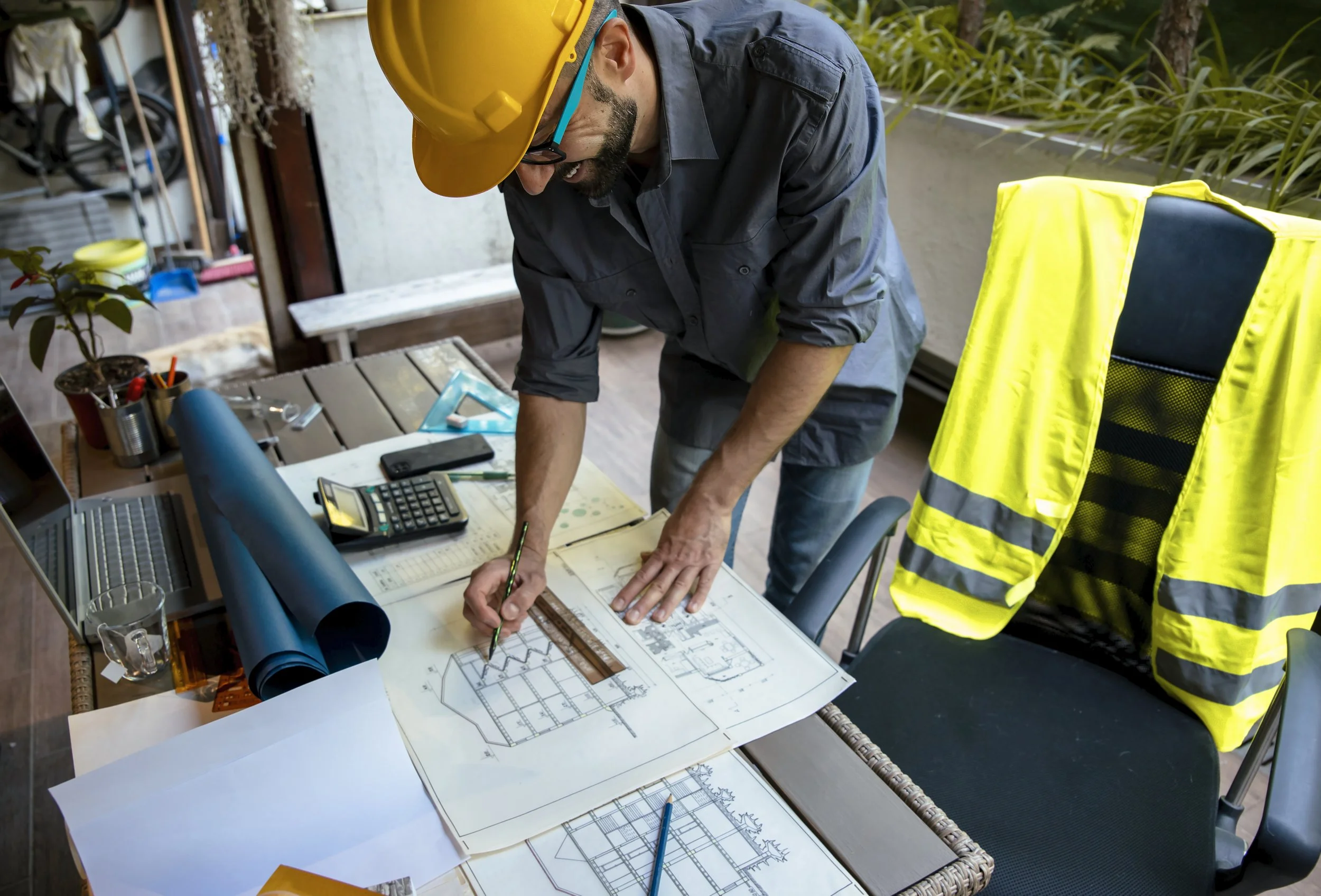 Un homme portant un casque de chantier travaille sur des plans architecturaux sur une table dans un bureau.