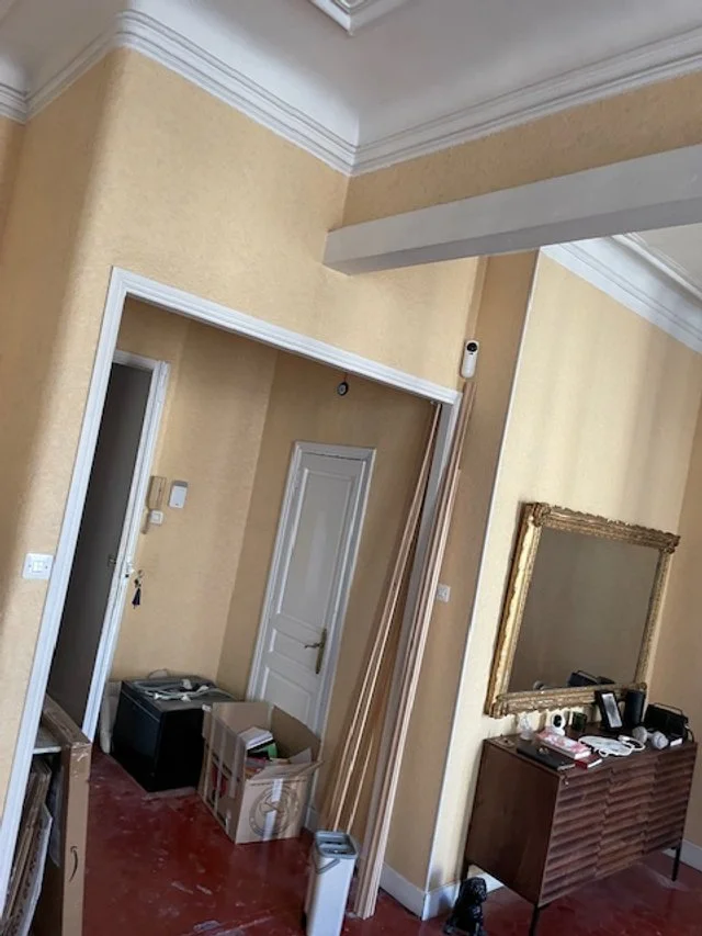 Intérieur d'un appartement ou d'une maison en rénovation avec un mur beige, une porte blanche, un miroir doré, une console en bois, et divers outils et cartons
