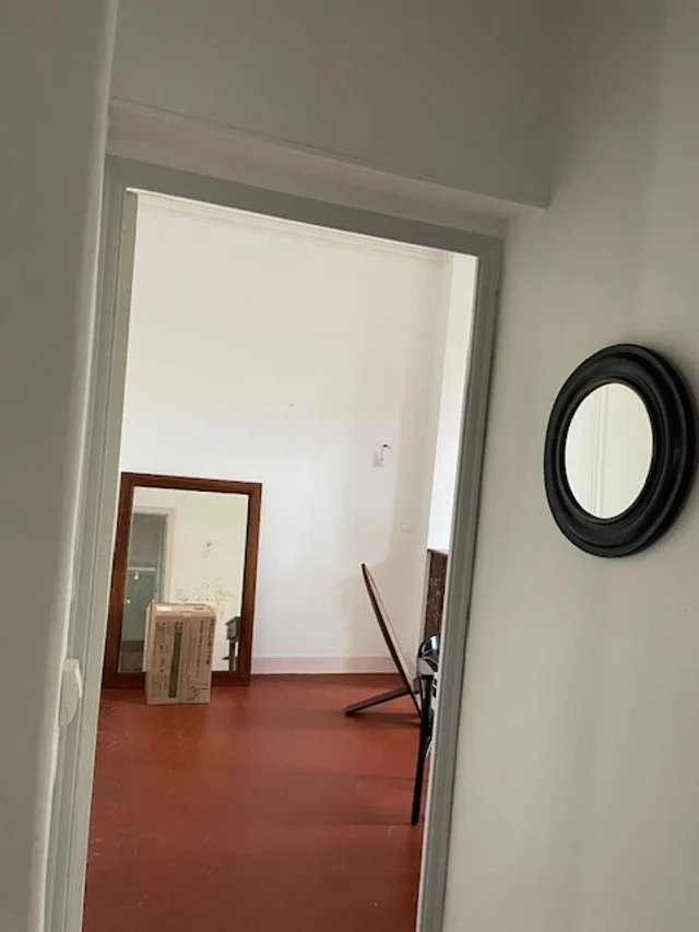 Une pièce avec un mur blanc, un miroir rond noir accroché au mur, et une porte donnant sur une autre pièce avec un plancher en bois, un cadre miroir posé au sol et une chaise renversée.
