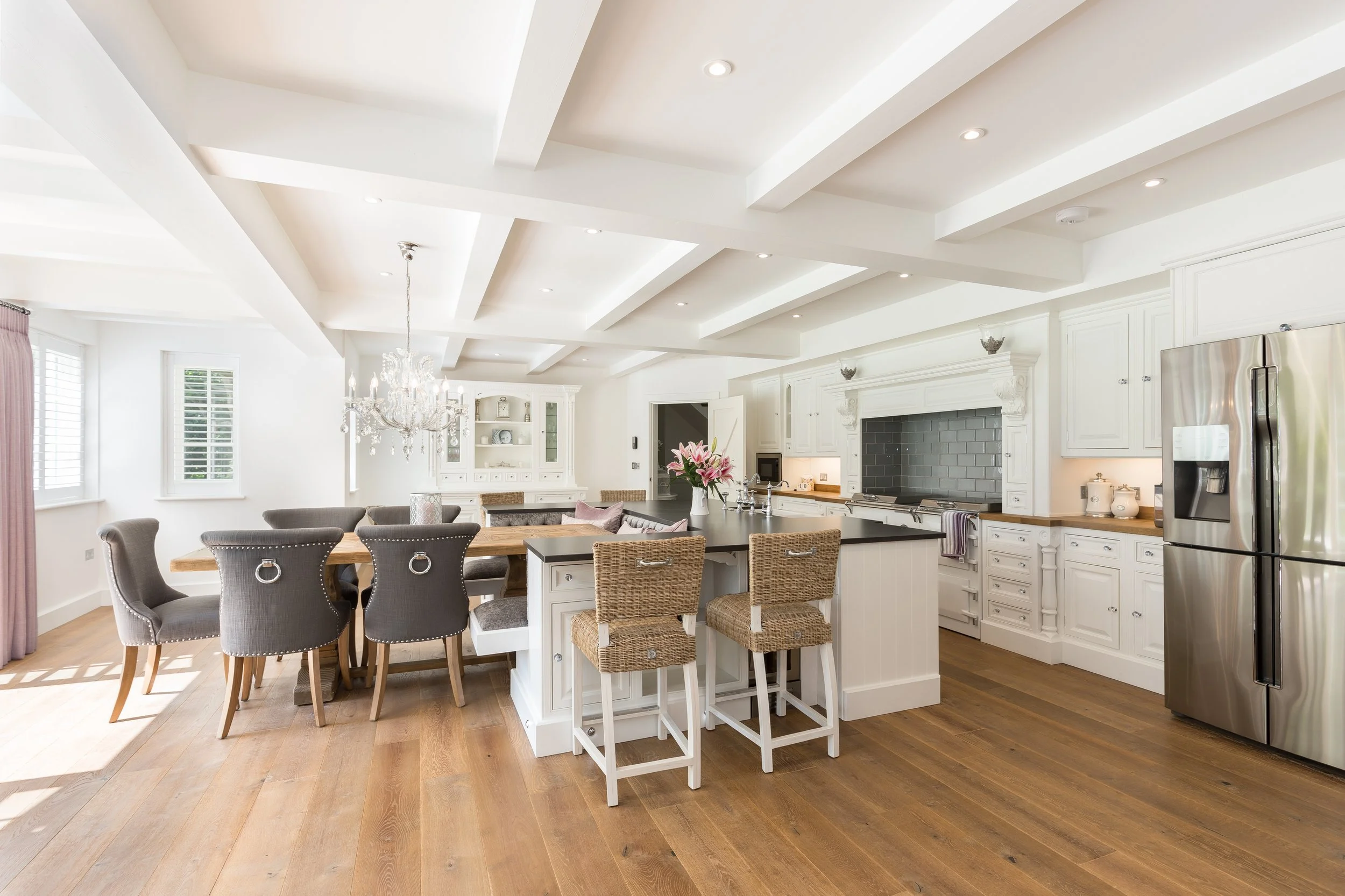 A spacious, bright kitchen with white cabinetry, a large central island with a black countertop, and a stainless steel refrigerator. There is a chandelier above the dining table on the left side of the image, which has six gray upholstered chairs aro