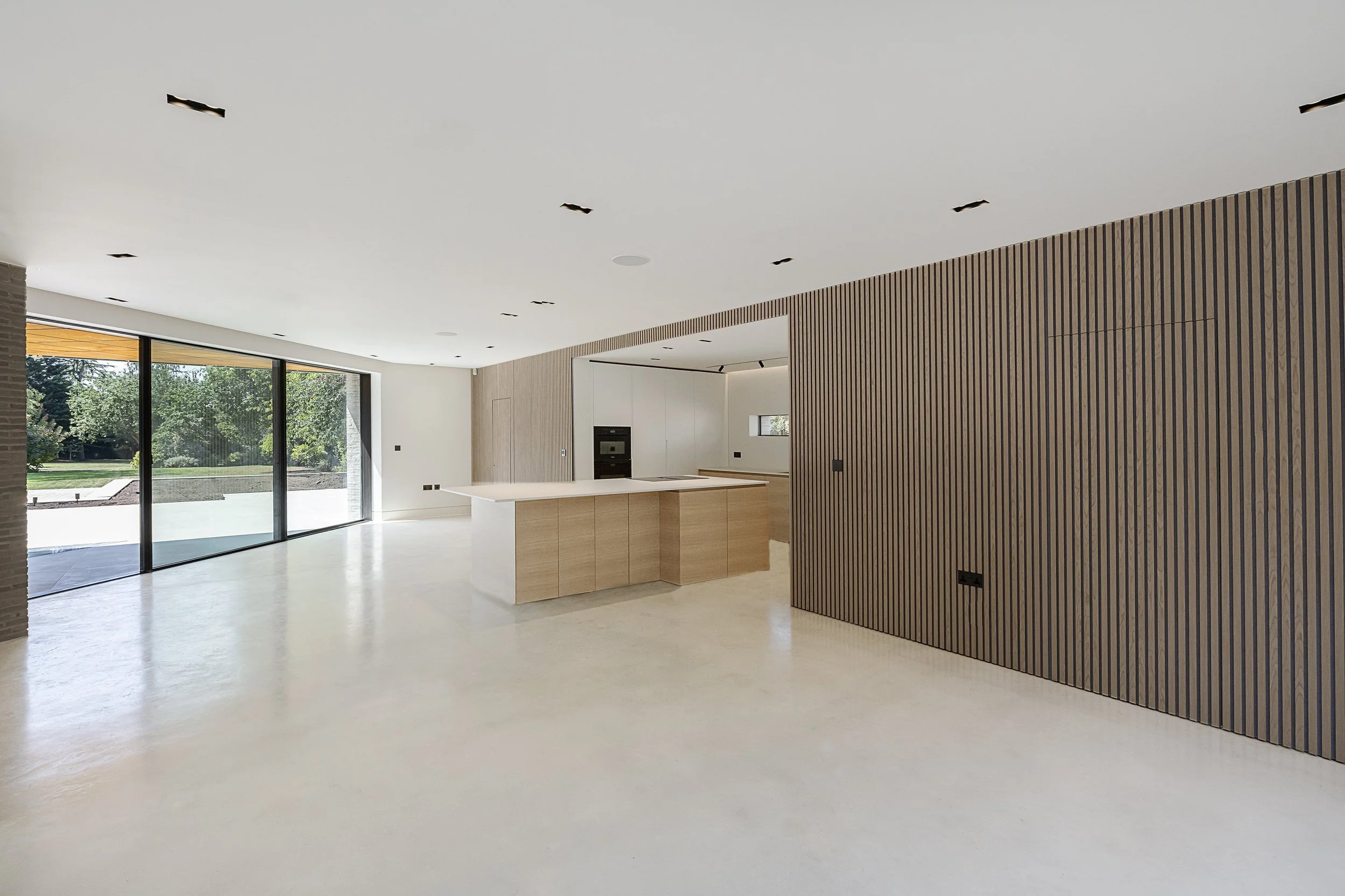 Modern, minimalist kitchen with white floor, large sliding glass doors, wooden accents, and an island with a light wood base and white countertop.