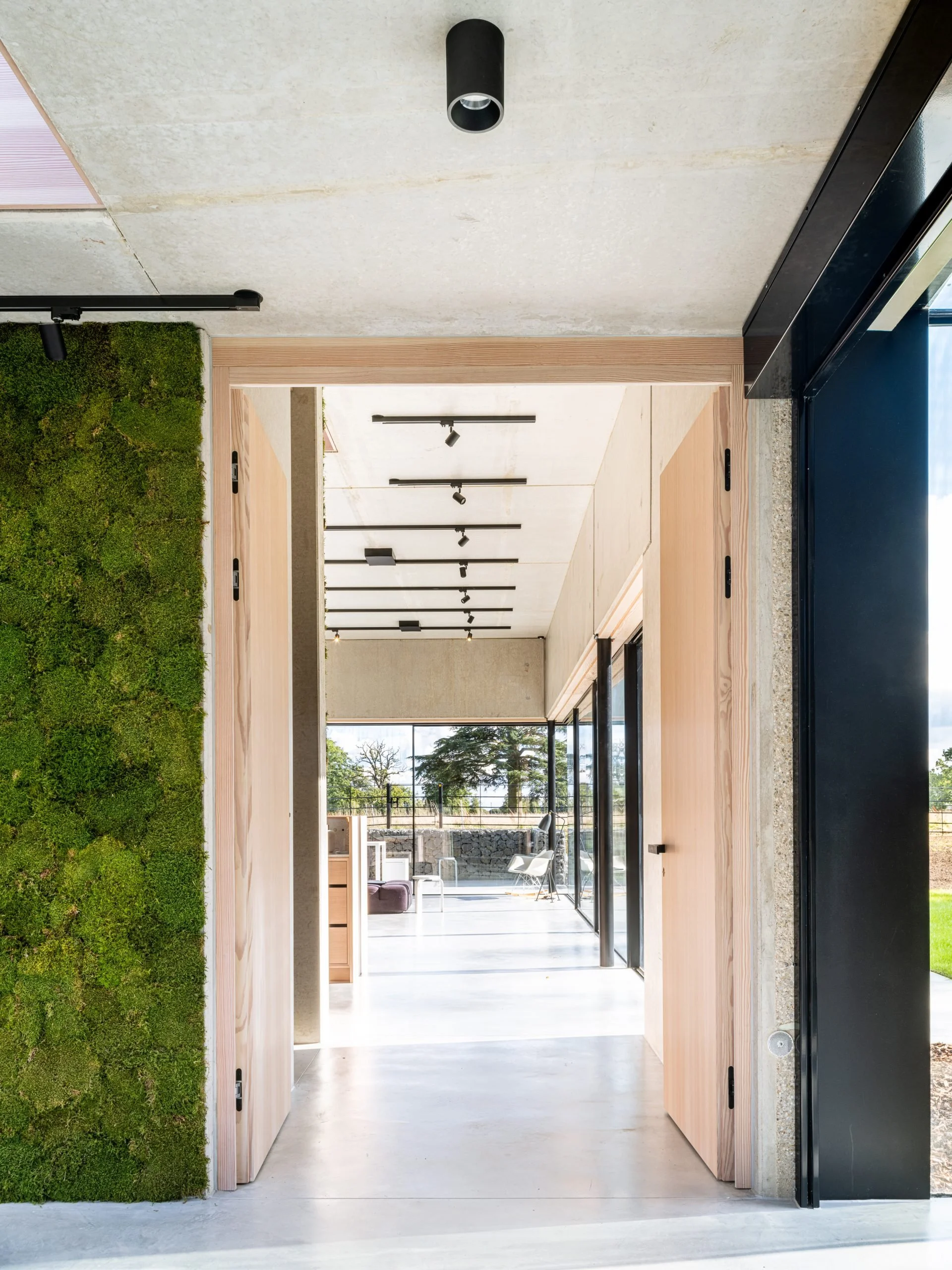 Modern interior space with sliding wooden doors, green moss wall on the left, concrete ceiling, and large glass doors at the back leading outside to a patio with chairs and trees.