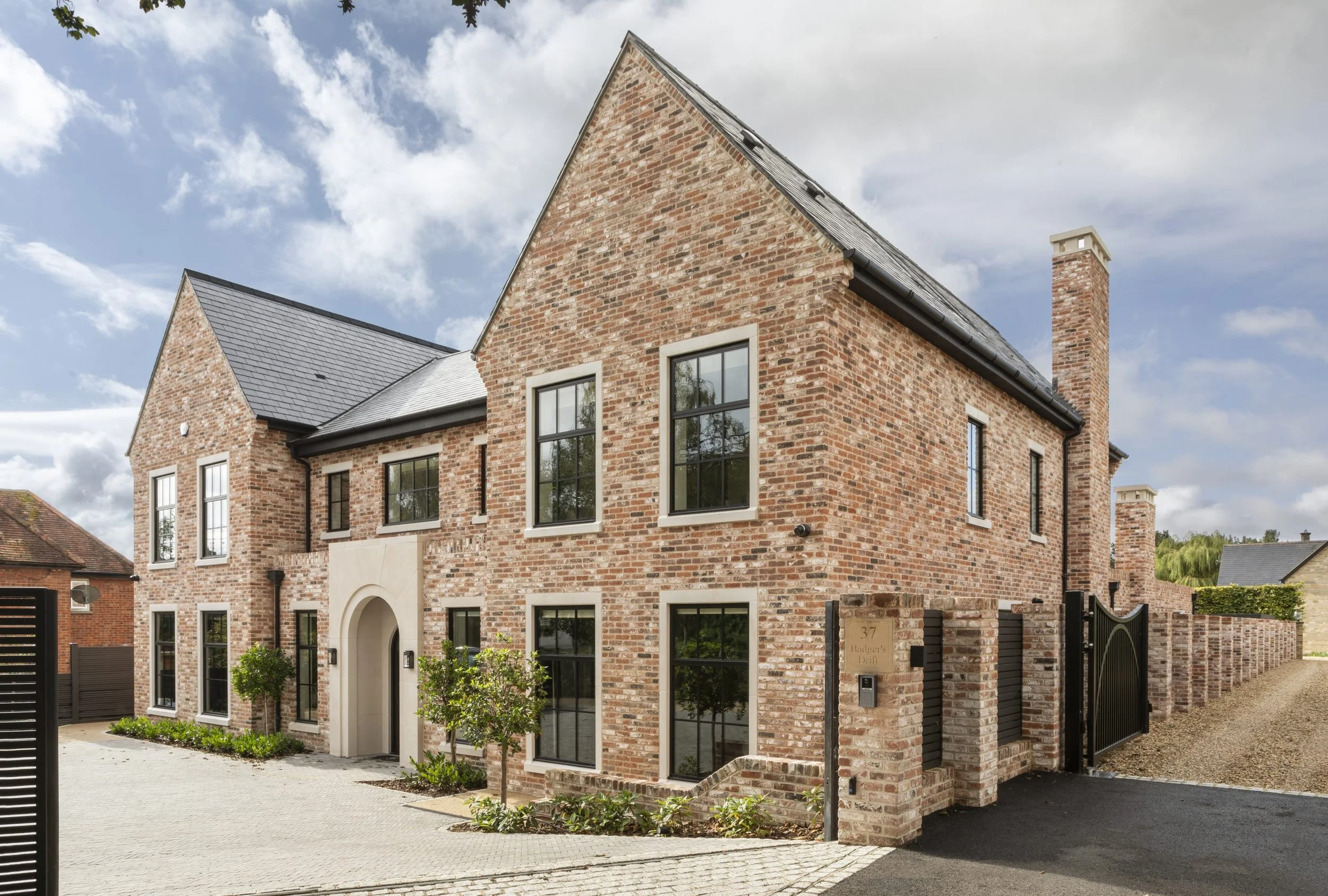 A brick house with multiple windows, black shutters, and a gated entrance. The house has a modern design with a sloped roof and chimney, surrounded by a paved driveway and small trees.