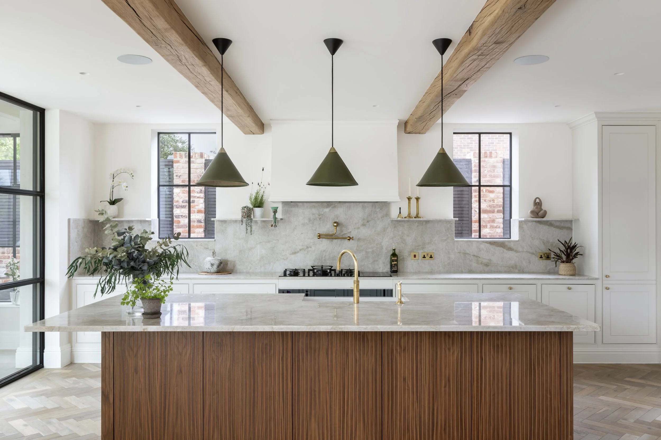 Modern kitchen with wooden ceiling beams, marble countertops, and pendant lights over an island with a brass faucet, decorated with plants and vases.