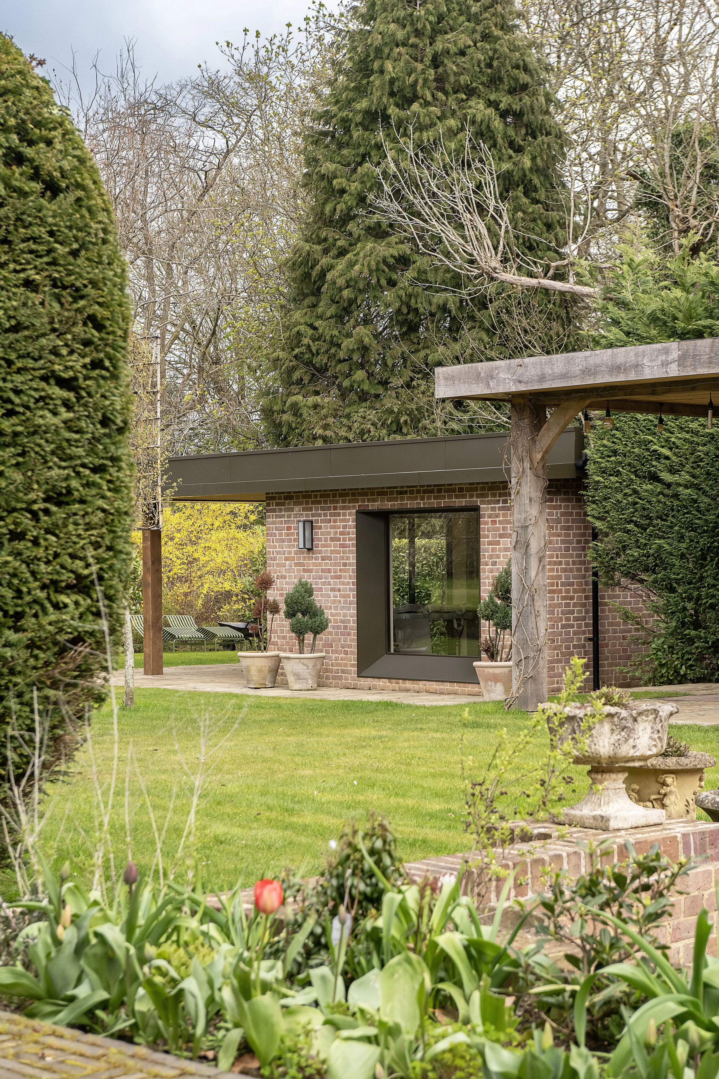A garden with green grass, potted plants, trees, and a brick building with a large window.