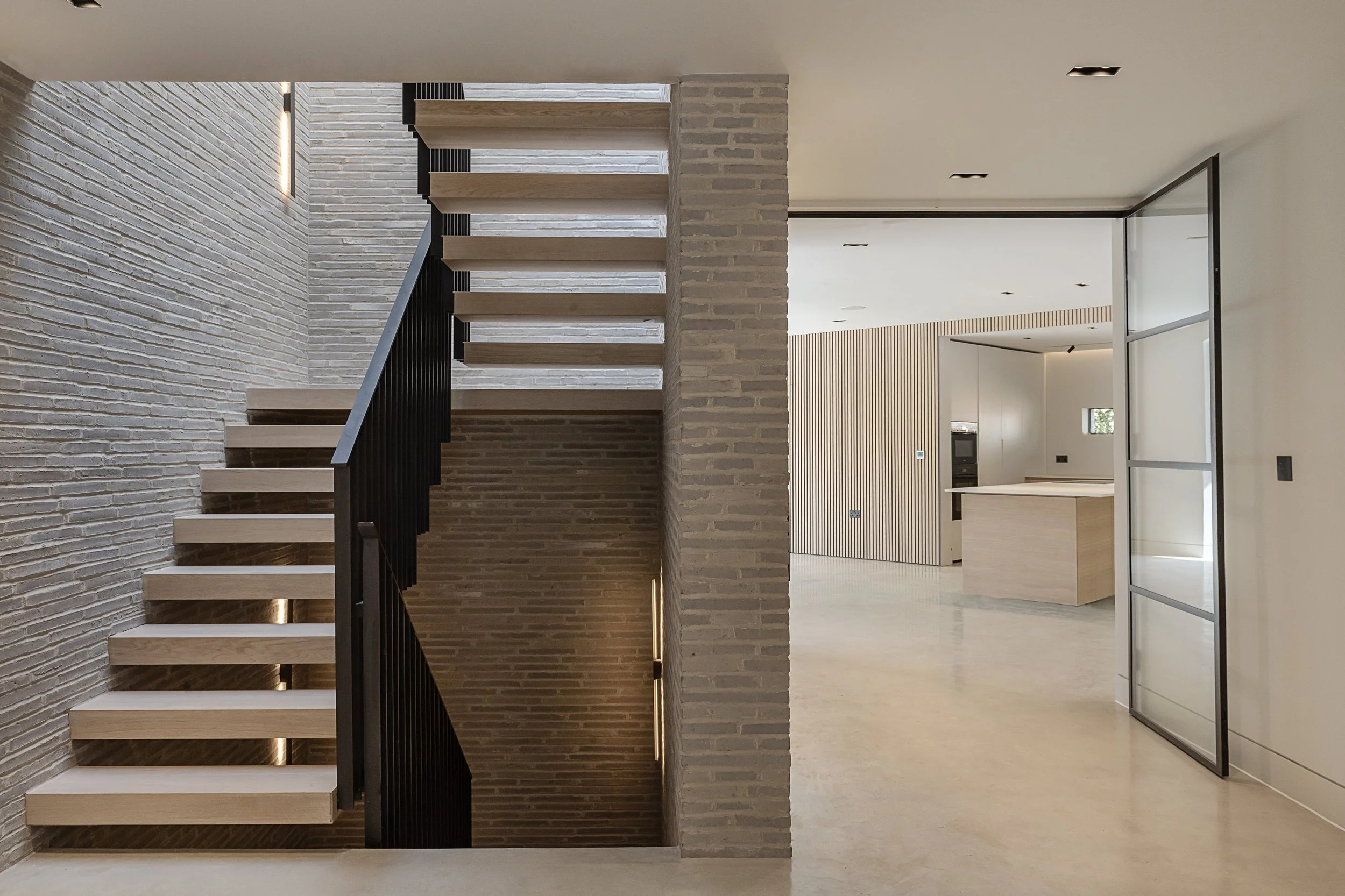 Modern staircase with wooden steps and black railing in a minimalist home interior with brick wall and open kitchen area.