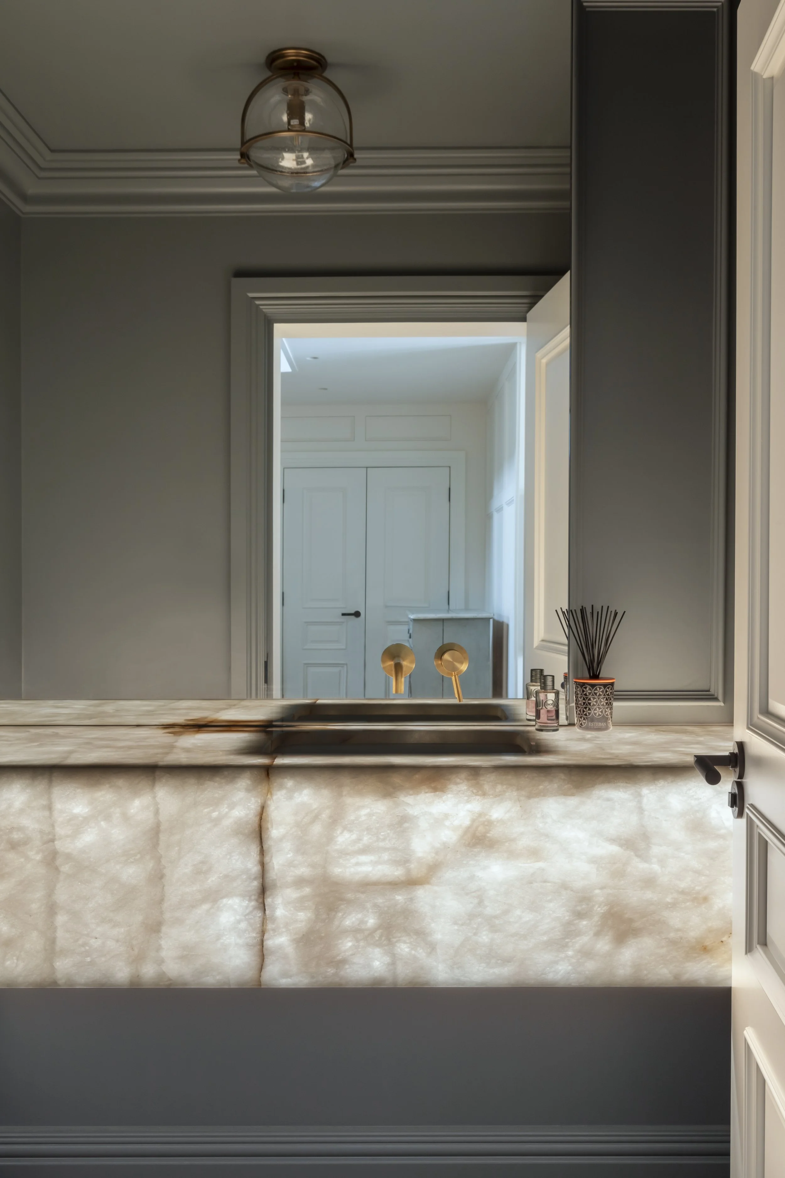 View of a modern bathroom with a marble countertop and sink, grey cabinets, and a large mirror. Visible doorways lead to other rooms, and a ceiling light fixture hangs overhead.
