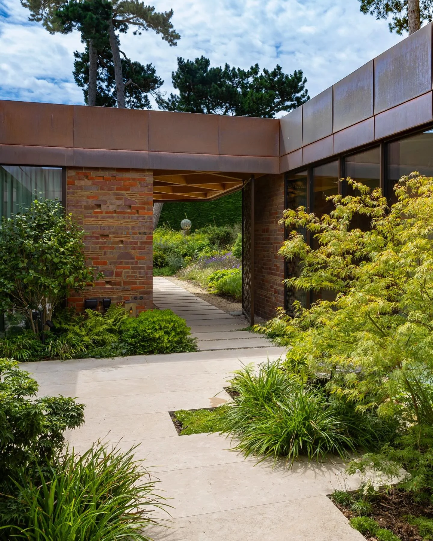 View of modern house entrance with a concrete pathway, lush green plants, brick walls, large windows, and trees in the background.