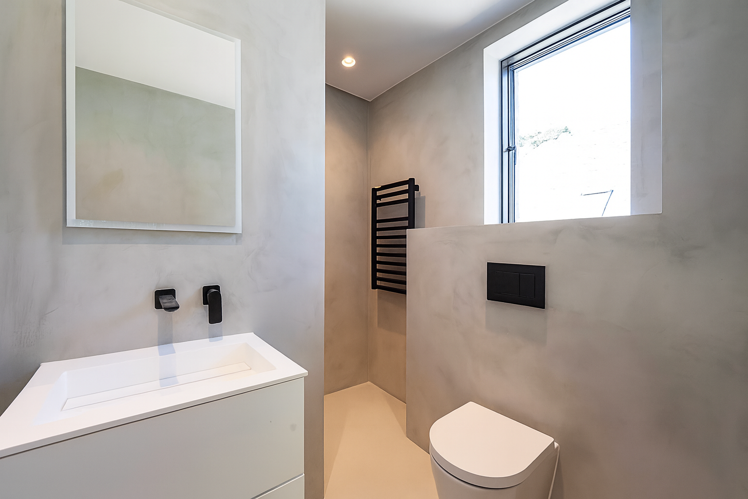 Modern bathroom with minimalist design featuring a white vanity with a integrated sink, black fixtures, a wall-mounted black towel warmer, a window bringing in natural light, and a wall-mounted black flush toilet.