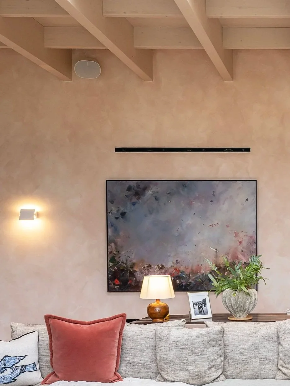 Living room with beige textured walls, a large sectional sofa with decorative pillows, a dark wood coffee table with candles and a tray, two table lamps, wall art, small vertical windows, a modern orange chair with a pillow, a white brick fireplace w