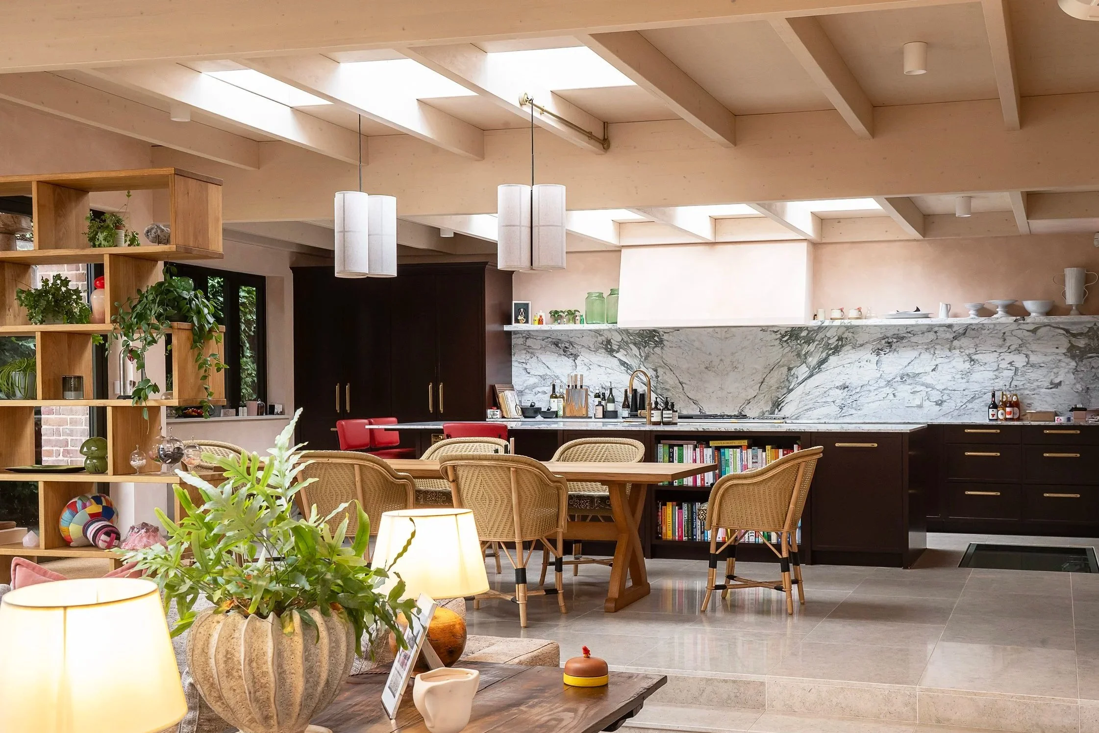 Open-concept kitchen and dining area with marble backsplash, black cabinets, a wooden dining table with five chairs, hanging pendant lights, and shelves with books and plants.