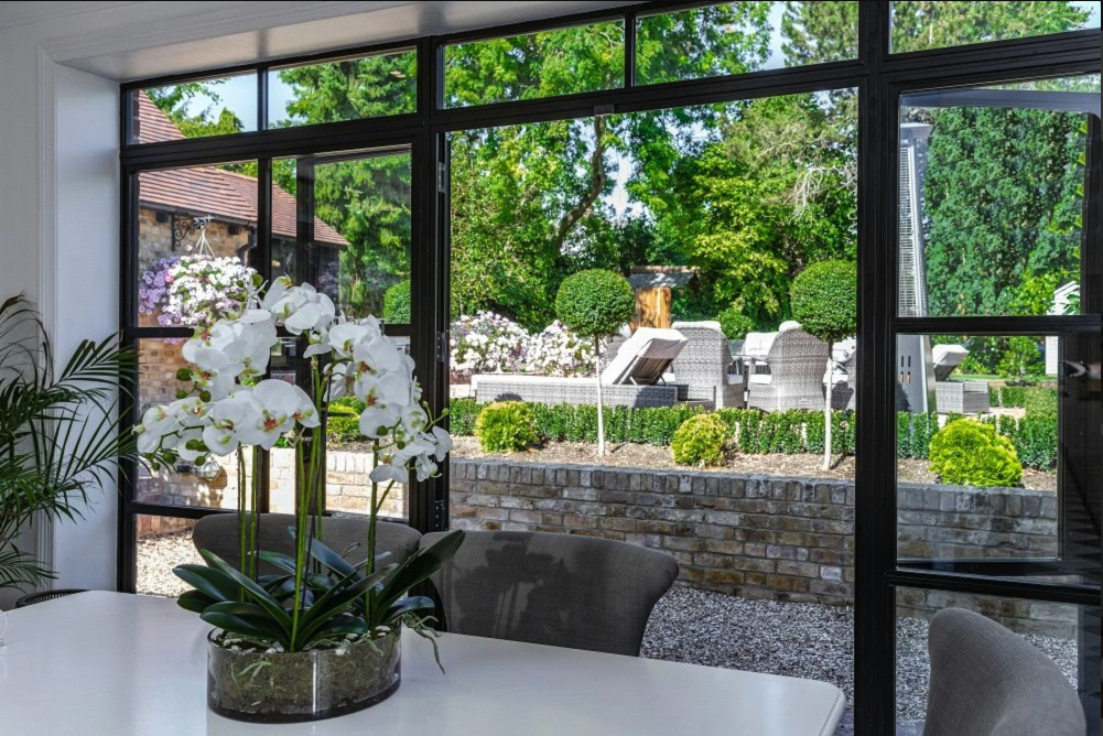 View from an indoor dining area looking out through large glass sliding doors to a landscaped backyard with outdoor seating, trees, and flowers.