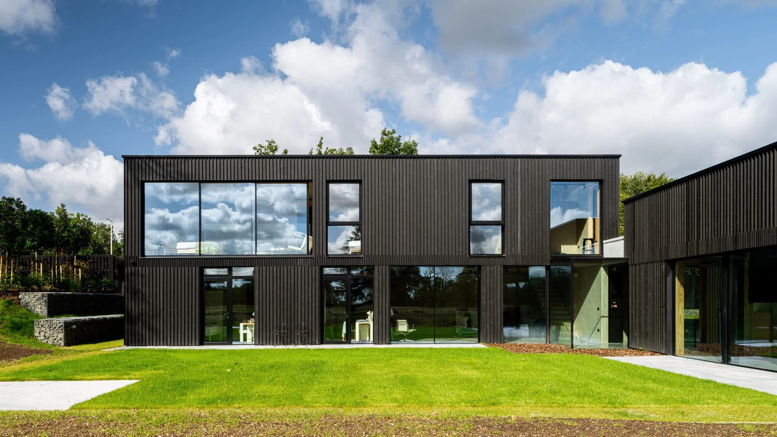 Modern two-story black building with large glass windows, green lawn in front, and partly cloudy sky.