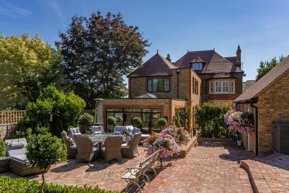 A backyard with a red brick patio, outdoor dining table with chairs, potted plants, flowering bushes, trees, and a large brick house with a steep roof and multiple windows.