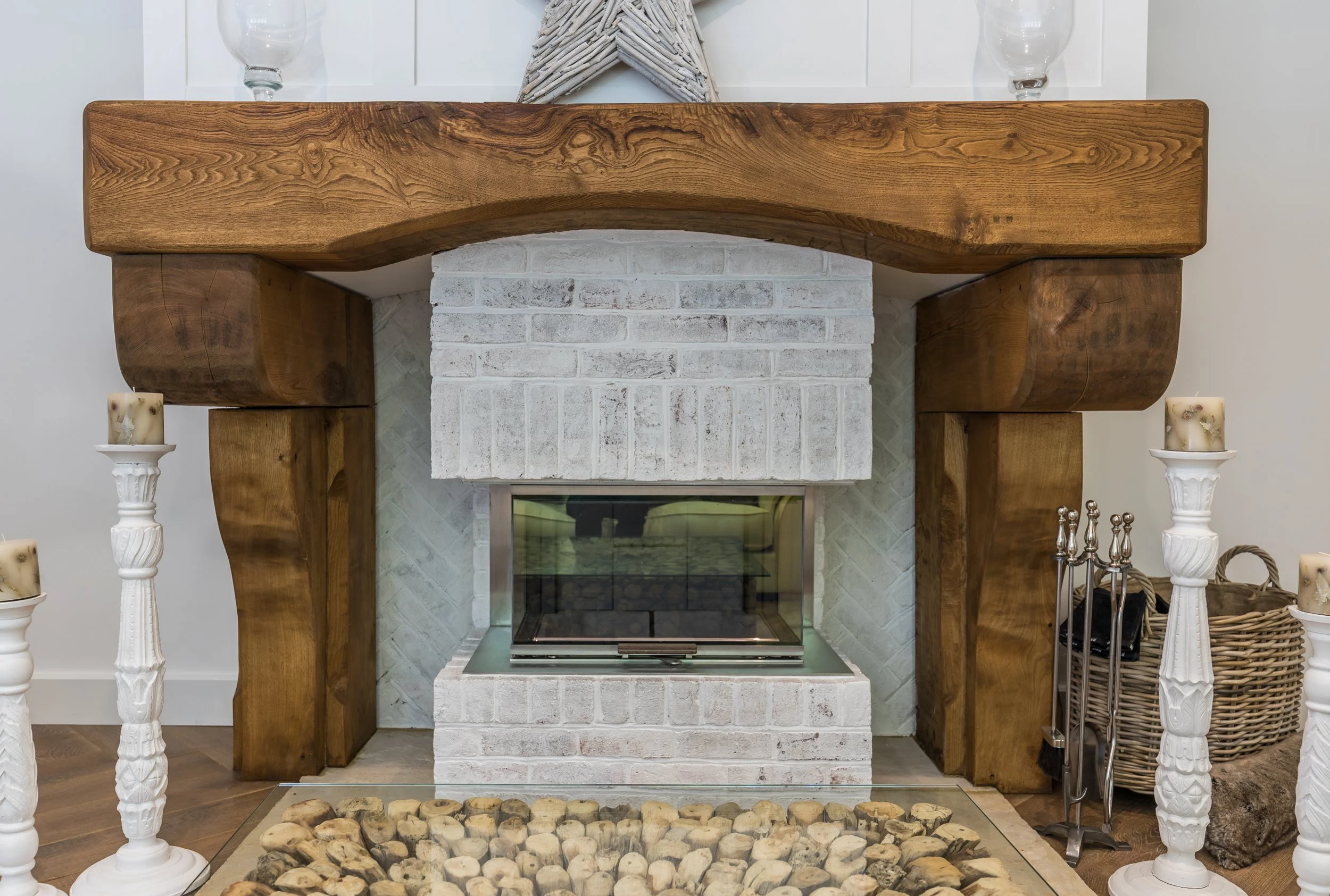 A white brick fireplace with a glass door, framed by large wood mantle and sides, with decor items including candles and a star on top, and a basket of fireplace tools to the right.