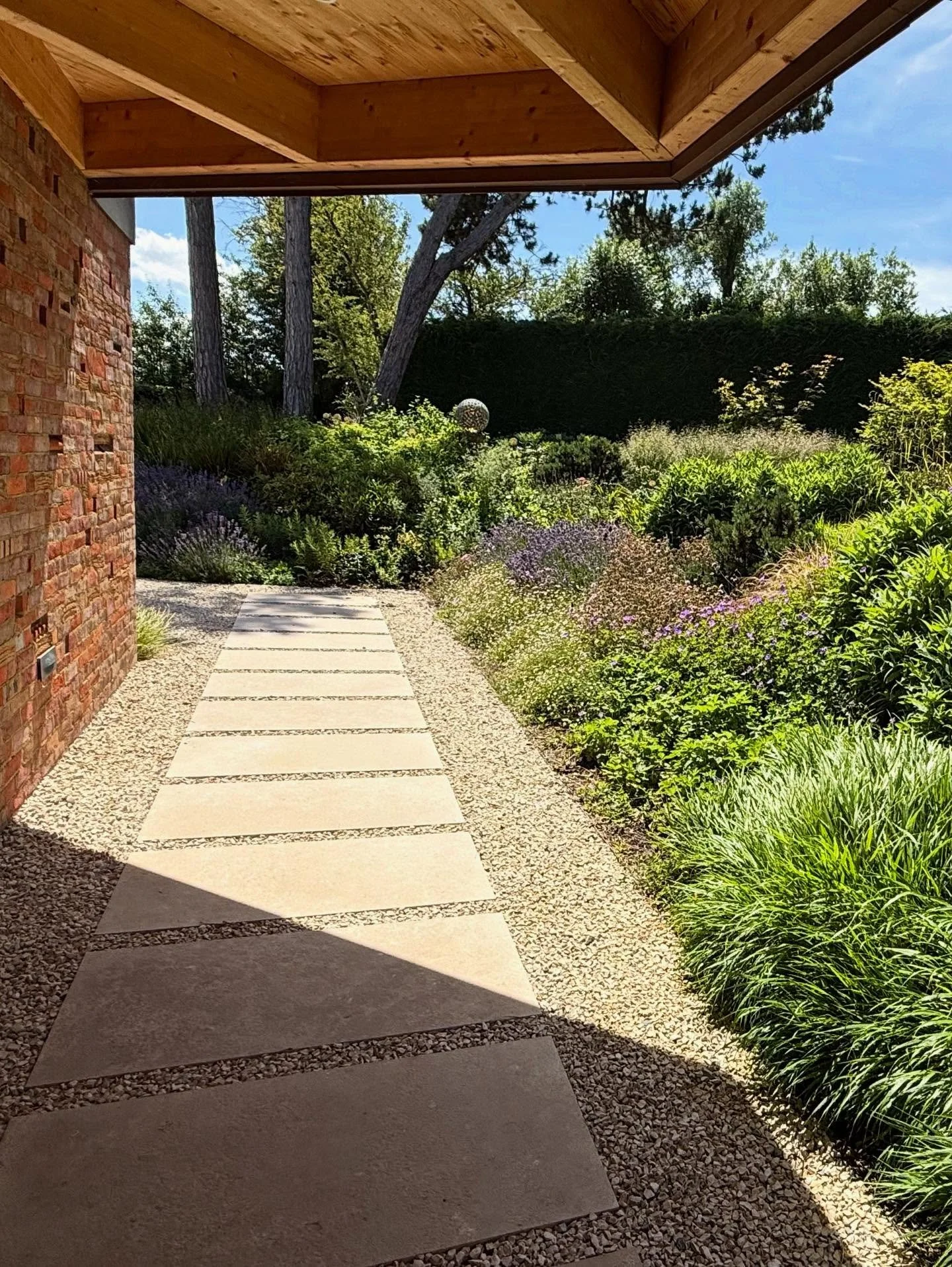 A garden pathway with large rectangular stepping stones leading away from a brick building, surrounded by lush green plants and purple flowers, with trees and a blue sky in the background.