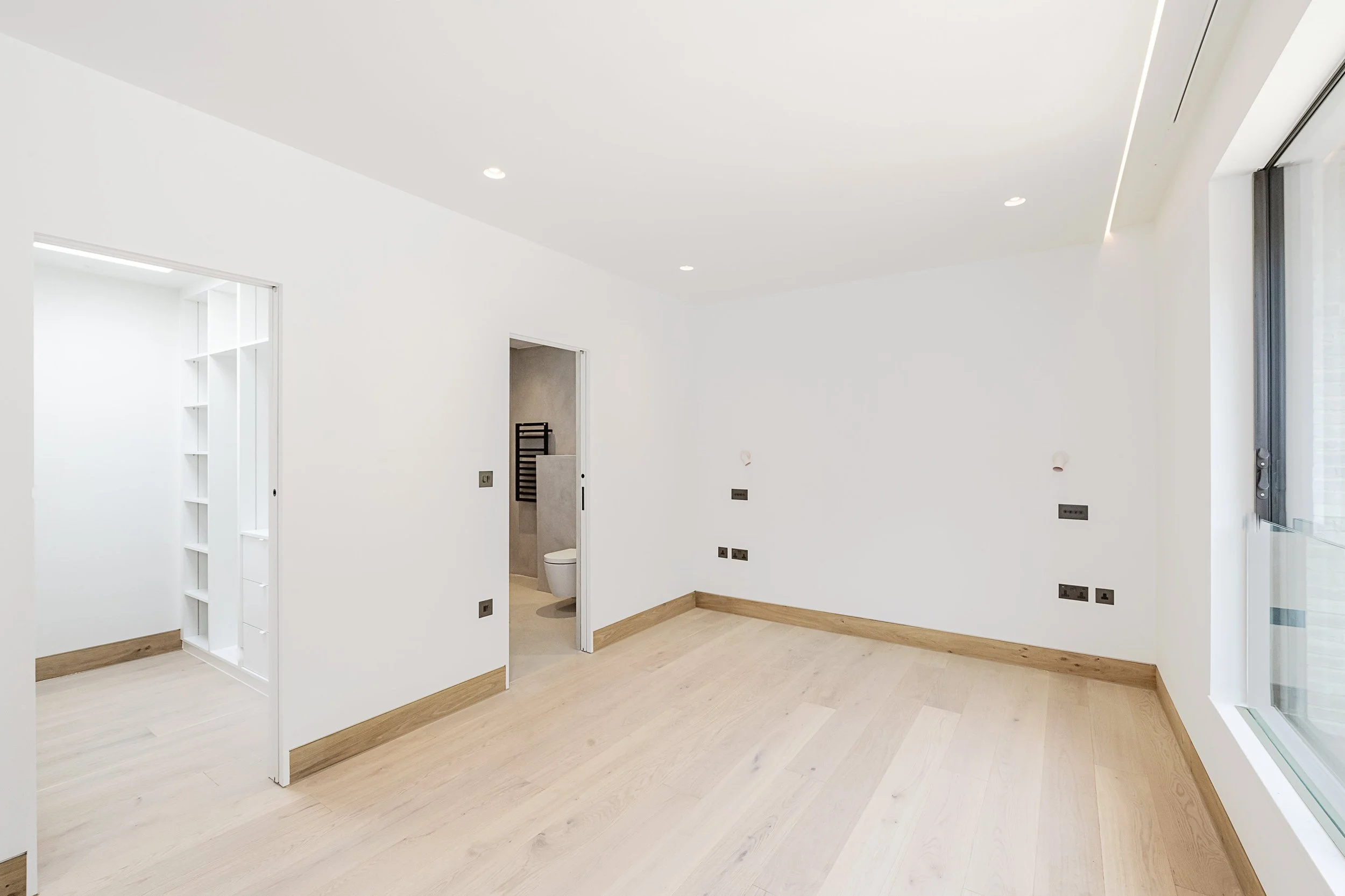 Empty room with white walls, light wood flooring, and a large window on the right. There is an open doorway to a small room or closet on the left and a bathroom with a toilet visible through a partially open door in the center.