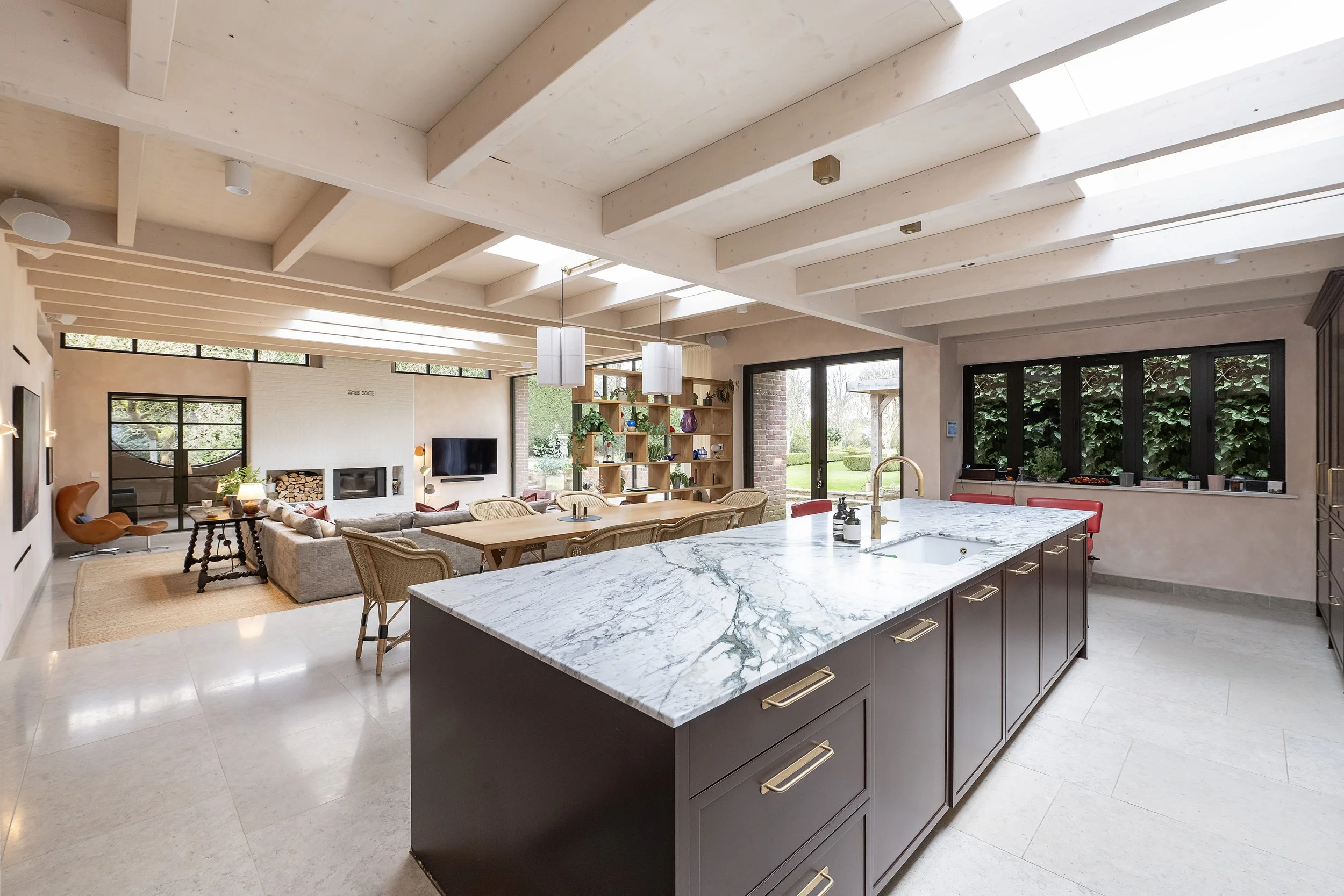 Open-concept living room and kitchen with marble island, beige walls, large windows, wooden beams ceiling, modern furniture, and outdoor view.
