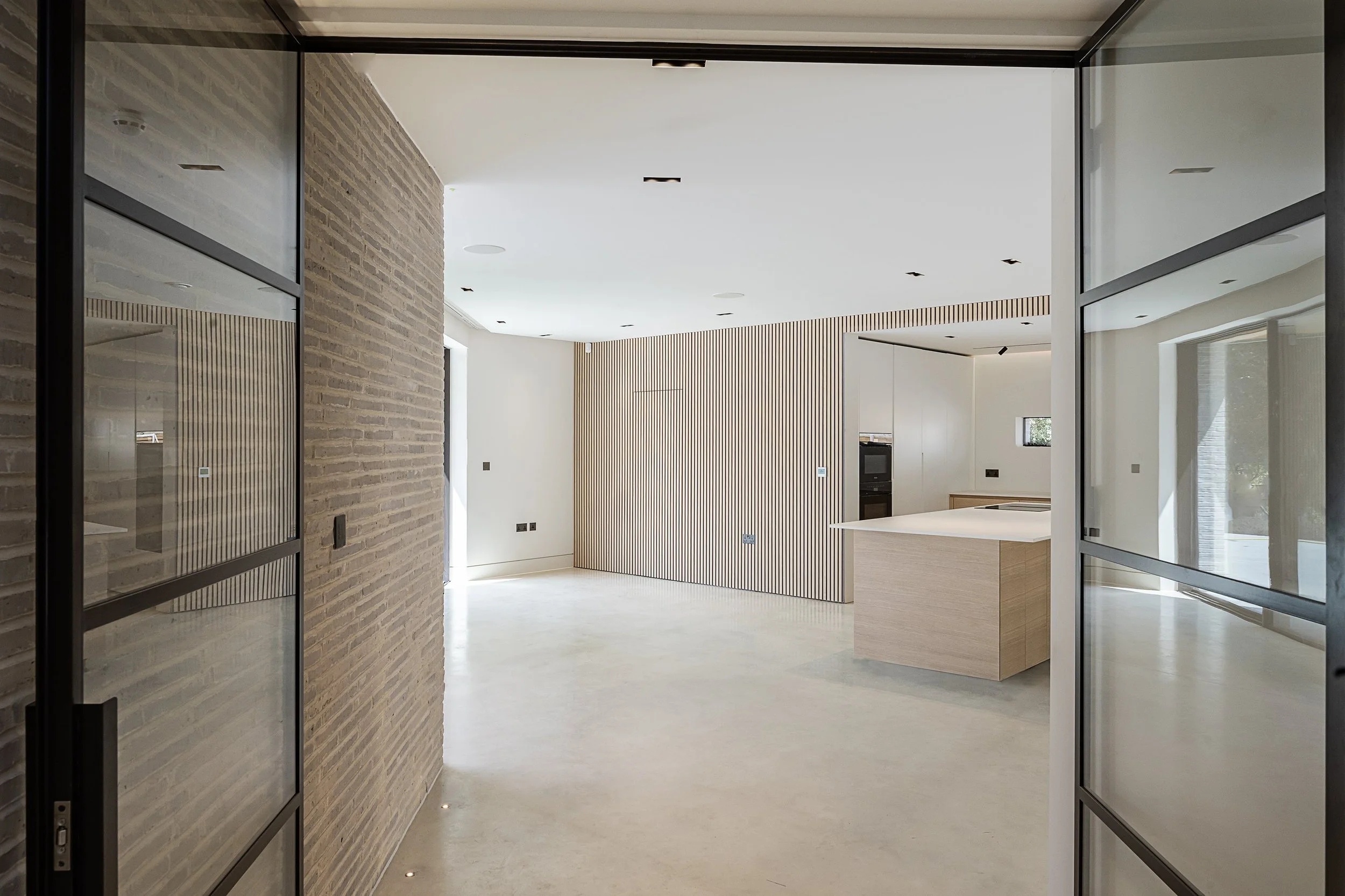 Modern, minimalist kitchen with light wood accents, white walls, and a curved wooden slat wall, seen through a glass door with black metal framing.