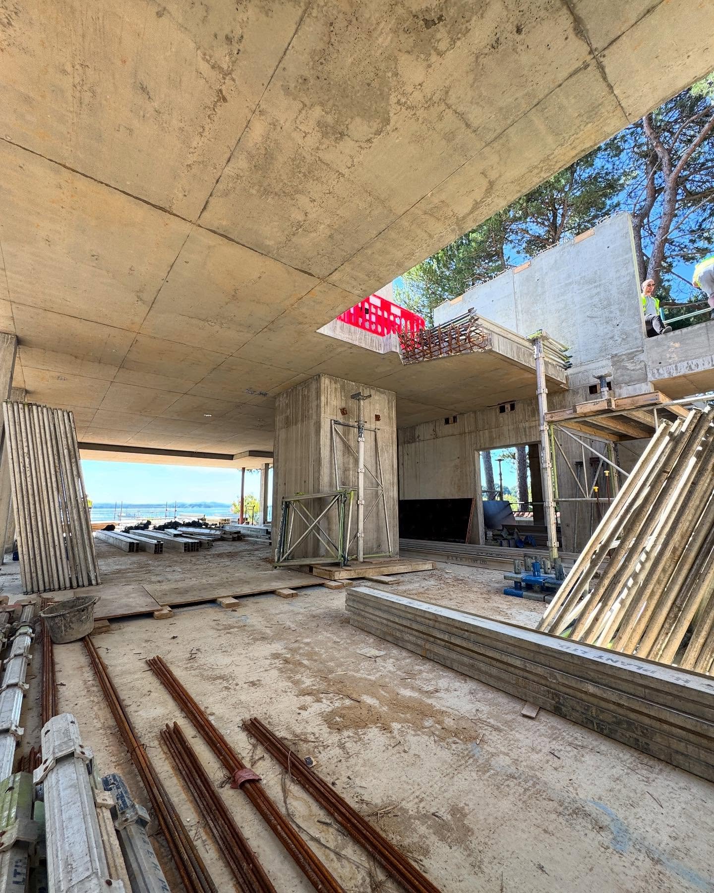 Construction site with concrete structures, metal rods, and scaffolding, open to the outside with trees and sky visible.