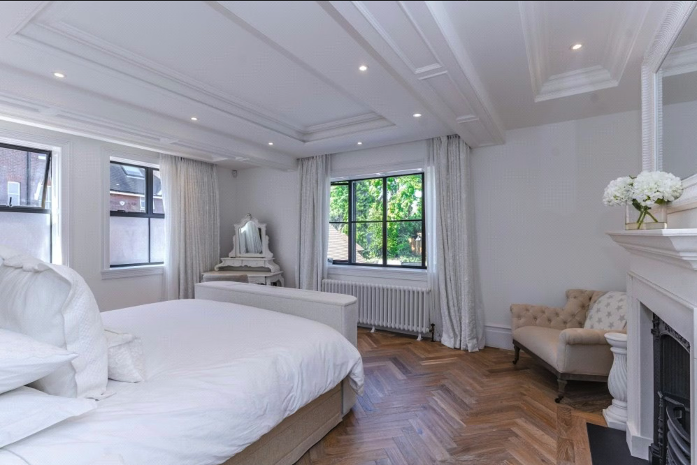 Bright bedroom with white walls, hardwood floors, a large bed with white linens, multiple windows with white curtains, a cream armchair, a white vanity with a mirror, and a fireplace with a floral arrangement.