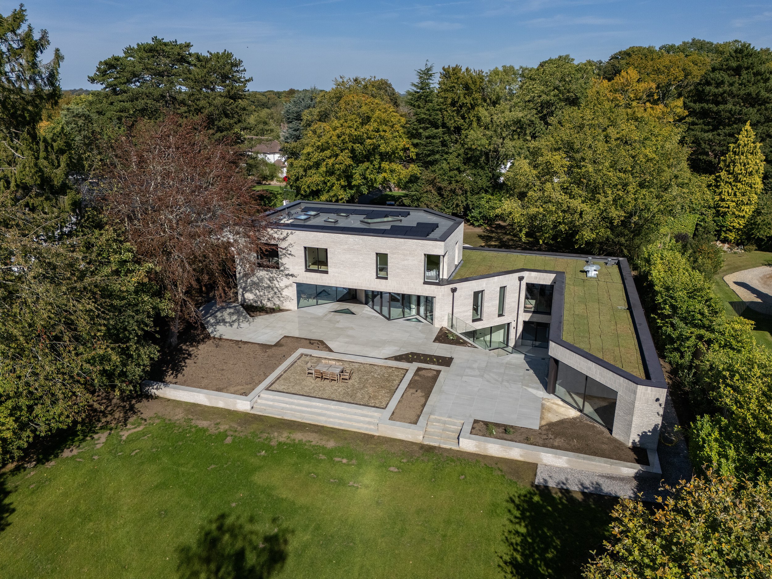 A modern house with a flat roof situated among tall trees, with a large outdoor patio and lawn.