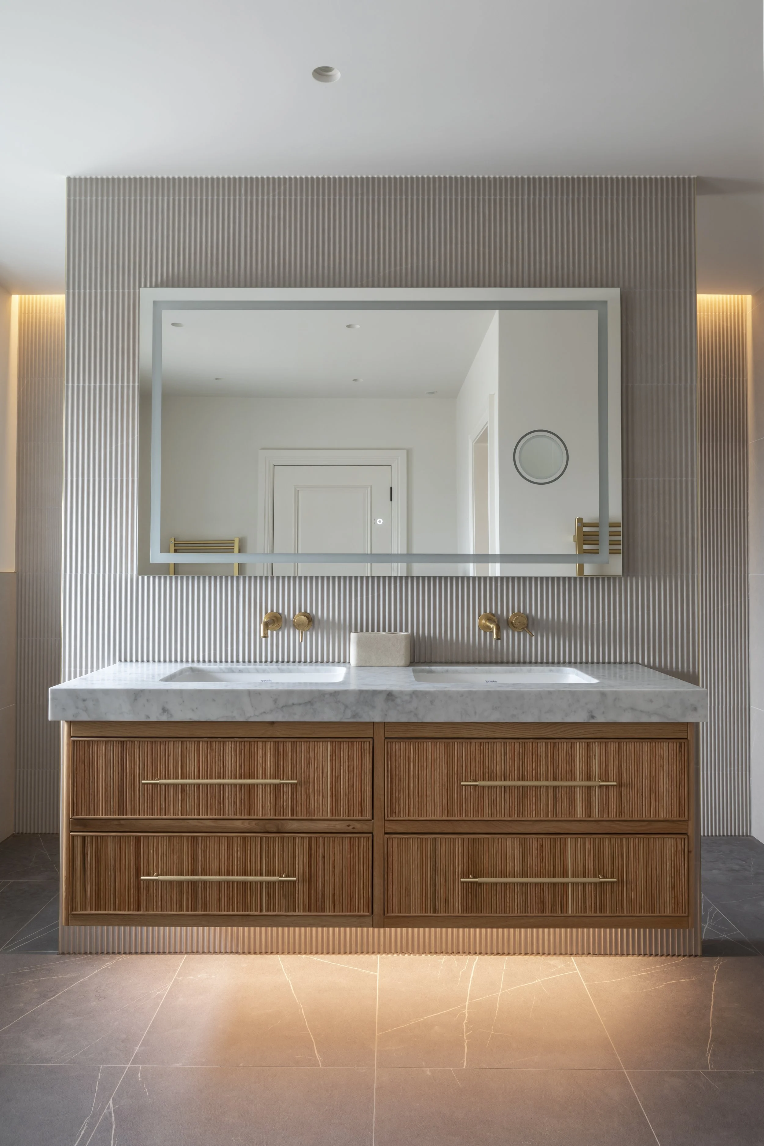 Modern bathroom with a double vanity, gold fixtures, large mirror, textured wall, and ambient lighting.