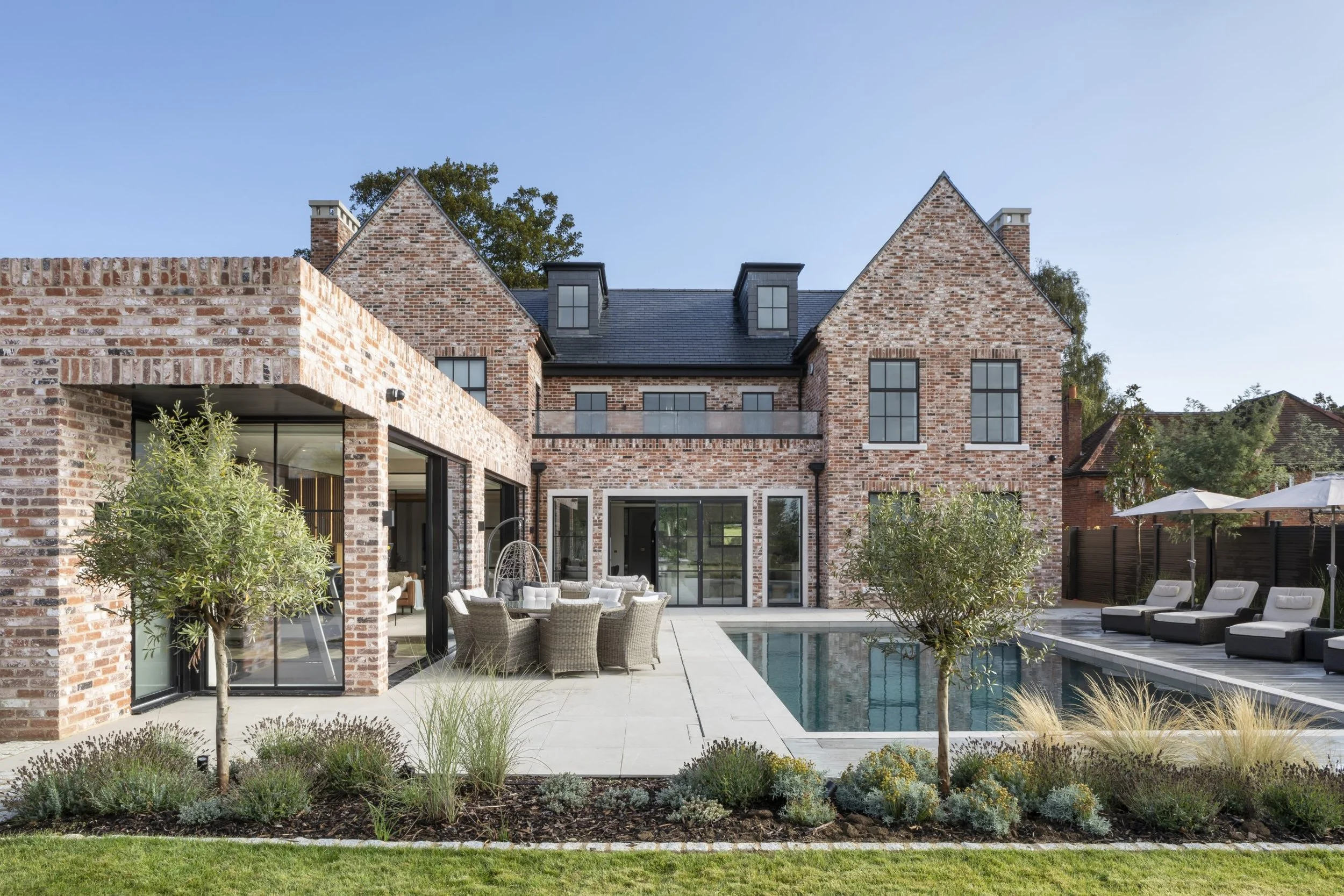 Brick house with a backyard pool, outdoor lounge chairs, patio furniture, and greenery.