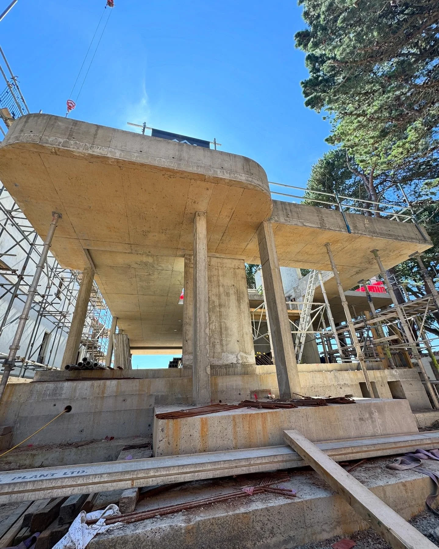 Construction site of a concrete building with scaffolding and support beams, overlooking a clear blue sky and tall trees.