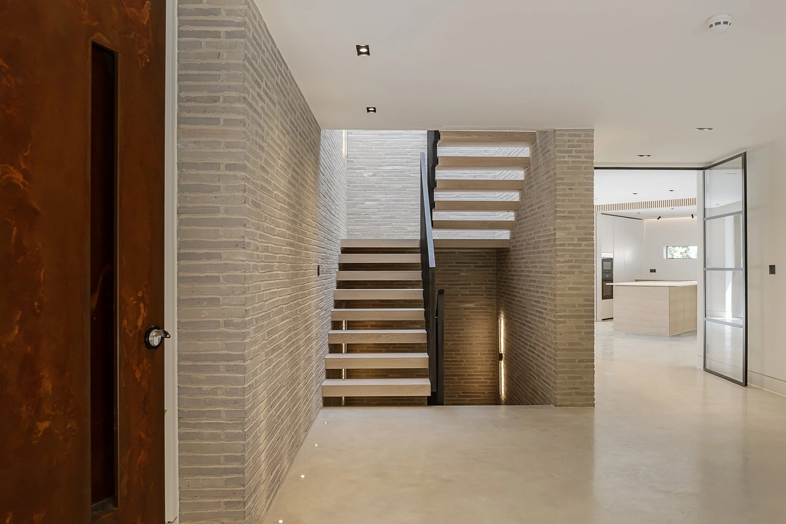 Modern interior with brick walls, a staircase with wooden steps and black railing, and a kitchen in the background.