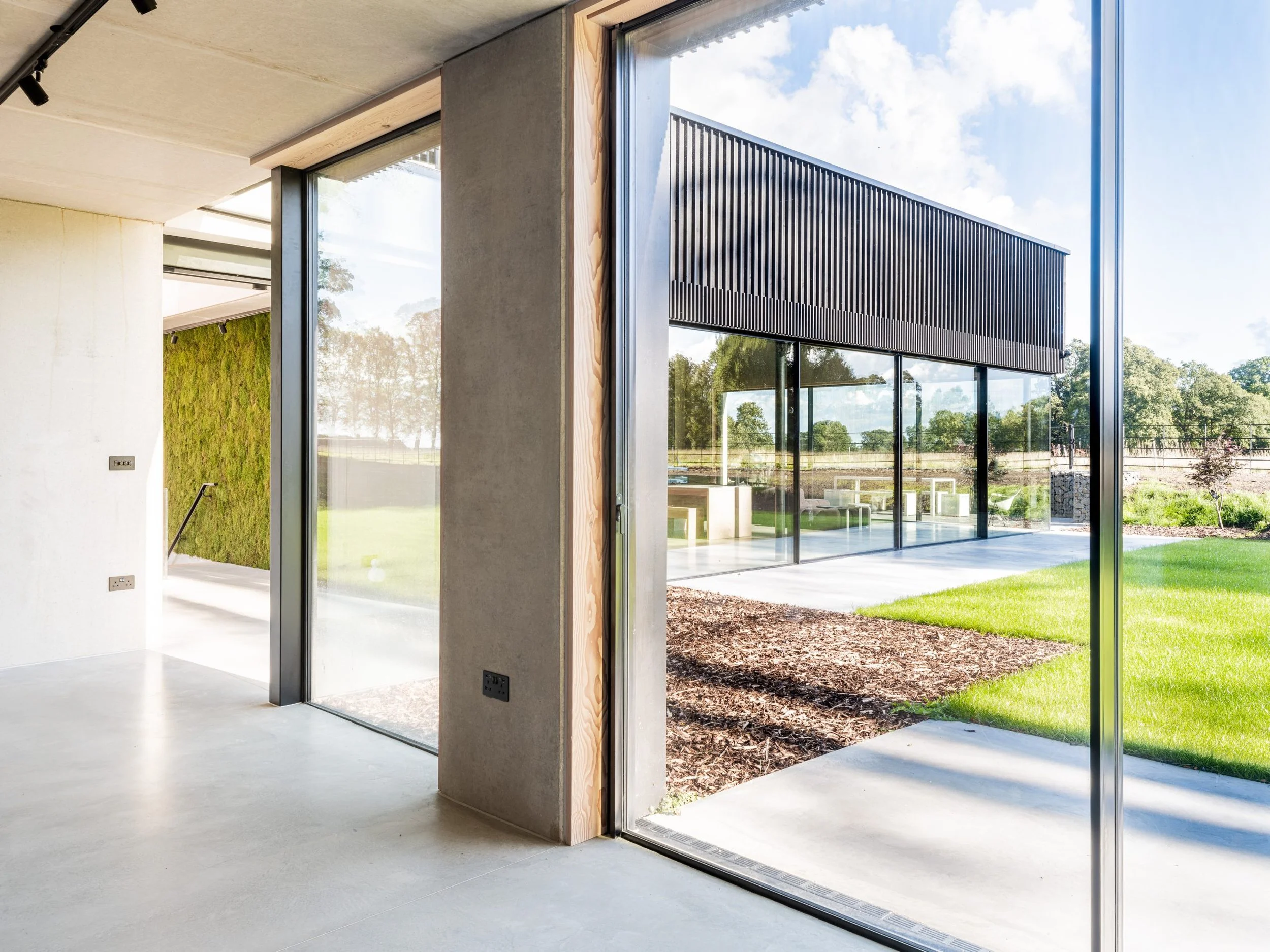 Modern interior space with large glass sliding doors opening to a landscaped yard with grass, trees, and outdoor furniture.