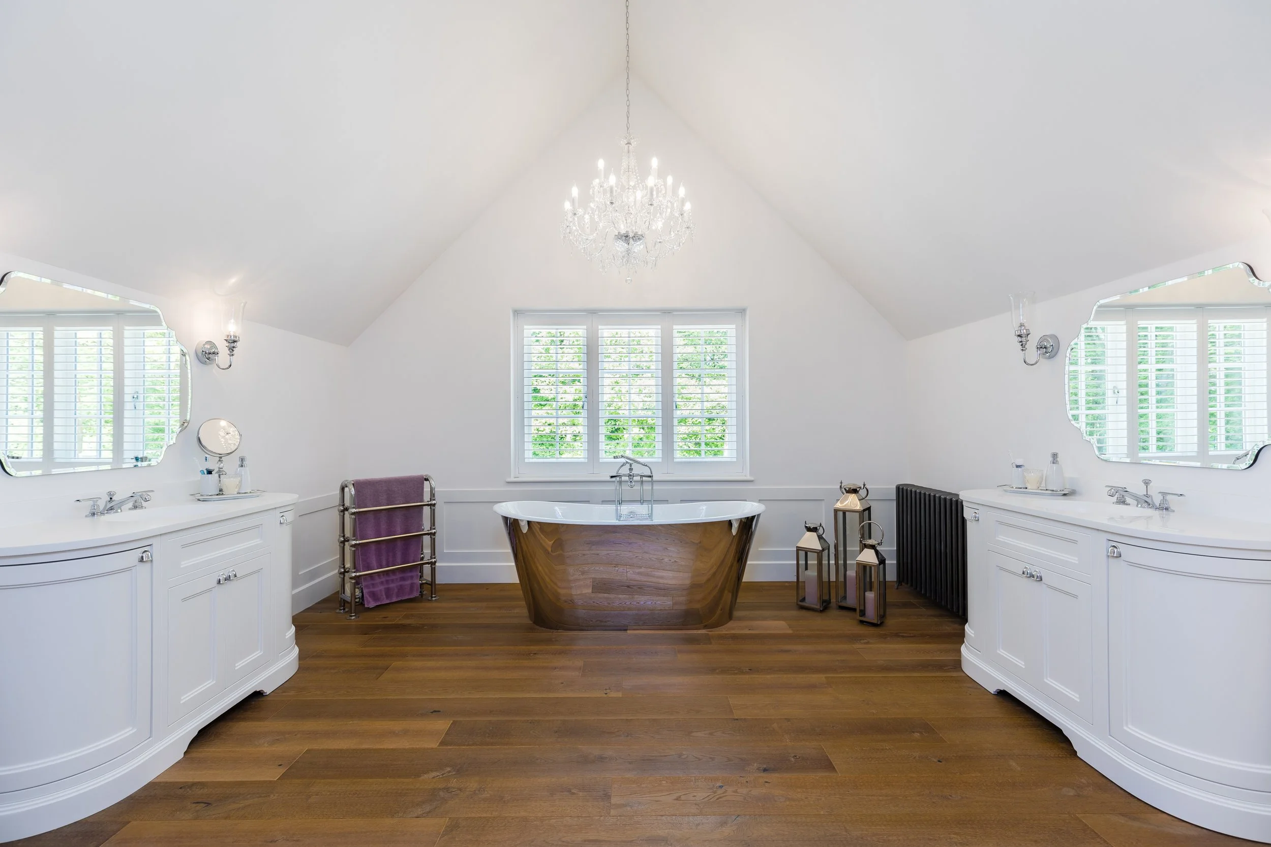 A bright, spacious bathroom with a central bathtub, white vanities on both sides, large windows with shutters, and decorative lanterns, chandelier, and wooden flooring.