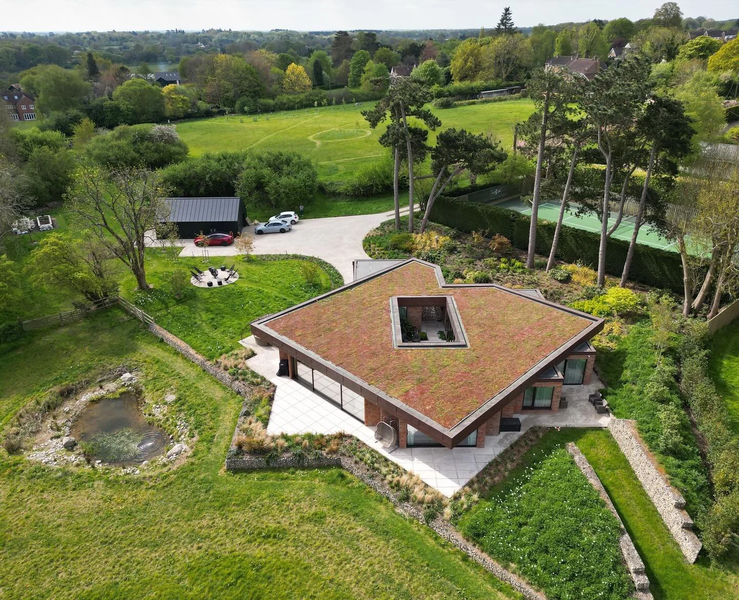Aerial view of a modern house with a green roof, surrounded by a garden with a pond, trees, and a patio. Tennis courts and a grassy field are visible in the background.