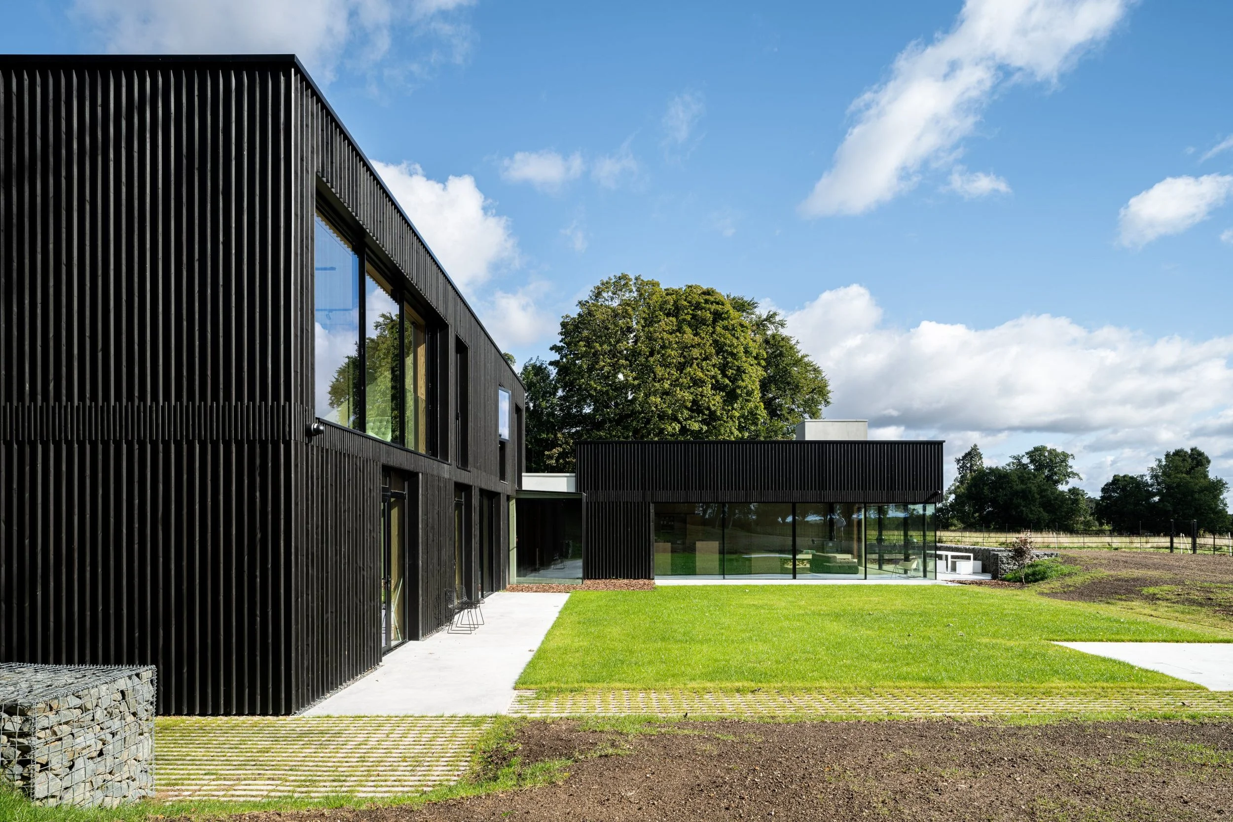 Modern house with black exterior walls, large windows, a green lawn, and trees in the background on a partly cloudy day.