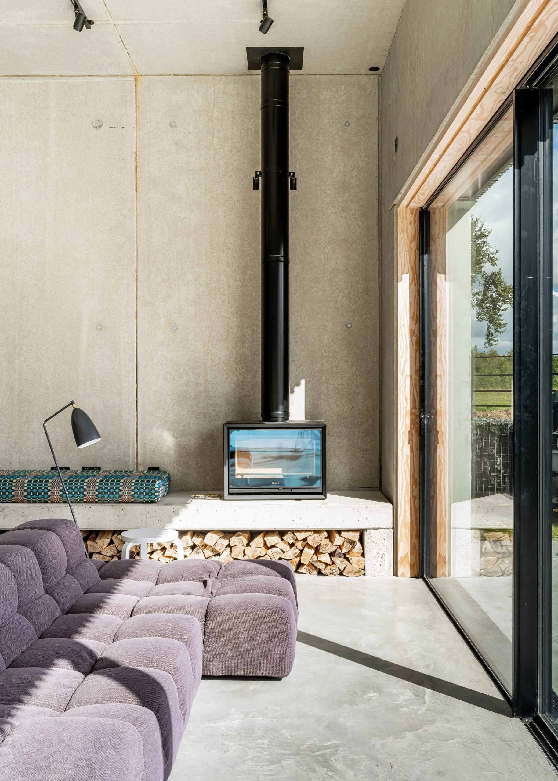 Modern living room with purple sectional sofa, concrete floor, wood and beige walls, a small white side table, a black floor lamp, a wood-burning stove with a glass door, and a large sliding glass door showing an outdoor landscape.