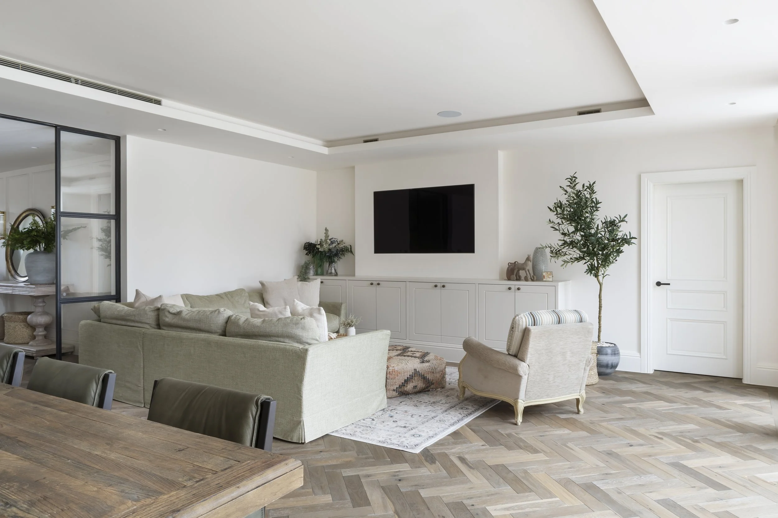 Bright living room with white walls, a beige couch, a vintage armchair, a large flat-screen TV on the wall, a white cabinet, a potted plant, and wooden flooring.