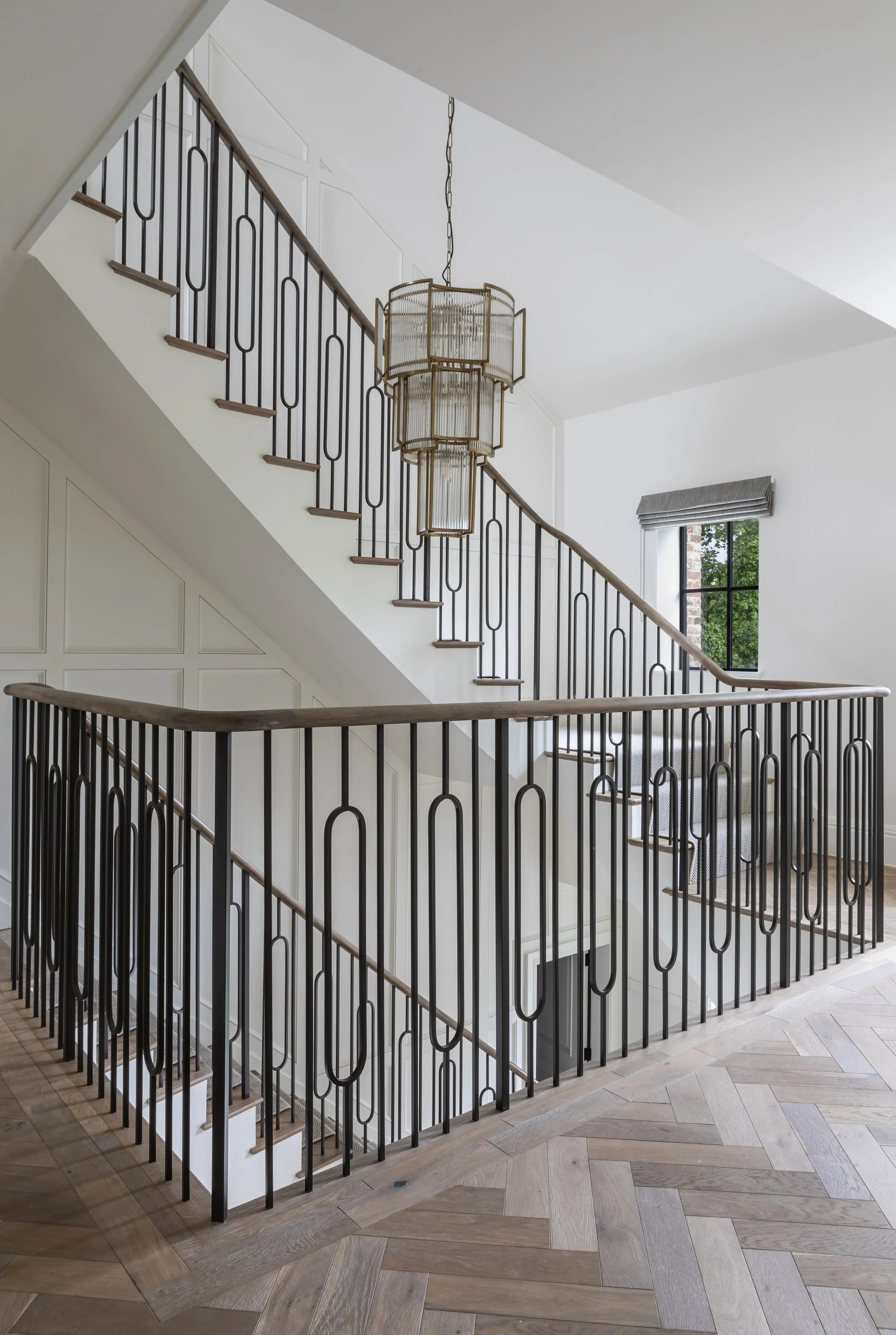 Interior view of a staircase with wooden handrails and black metal balusters, a large multi-tiered gold chandelier hanging from the ceiling, a window with a gray Roman shade, and wood flooring arranged in a herringbone pattern.