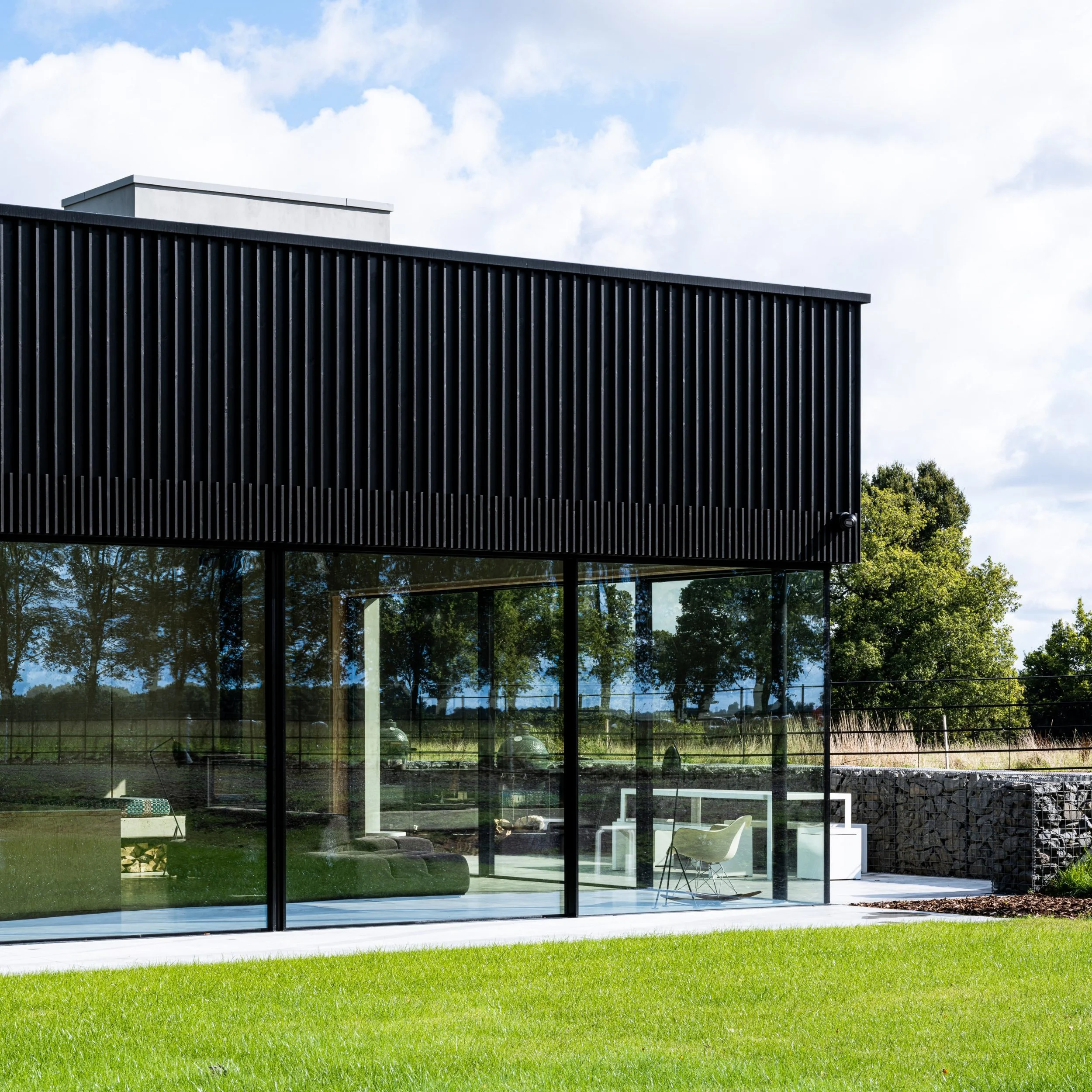 Modern building with black exterior and large glass windows, surrounded by green lawn and trees, under a partly cloudy sky.